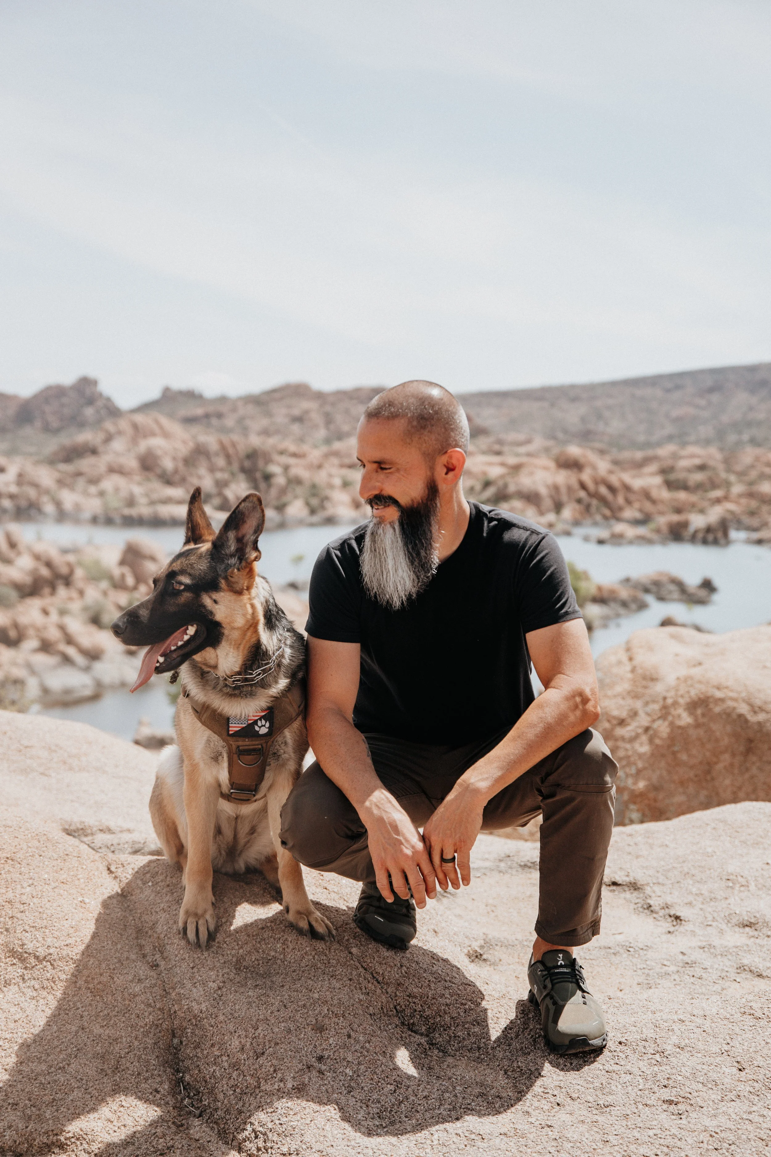 A man with a beard and a shaved head, dressed in a black t-shirt and brown pants, crouching on rock, smiling at a German Shepherd dog sitting beside him, in a rocky desert landscape with a body of water in the background.
