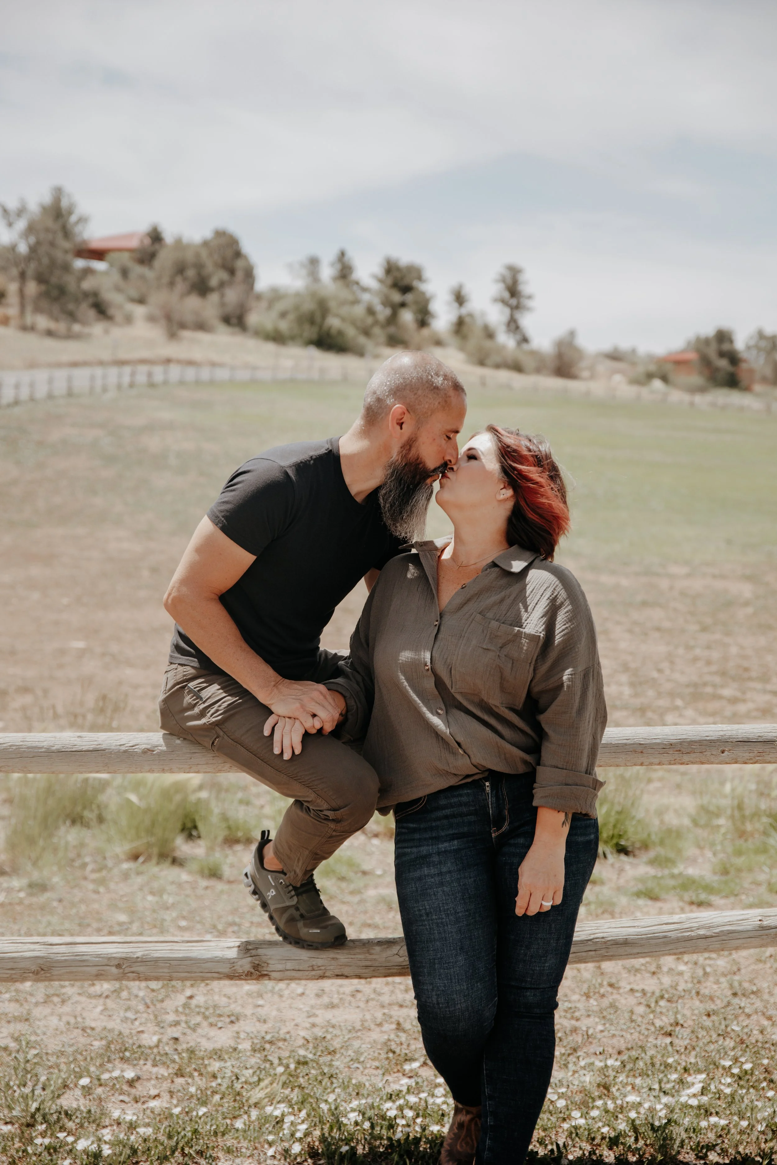 A man with a beard and a woman with red hair exchanging a gentle kiss outdoors near a wooden fence with a grassy field and trees in the background.