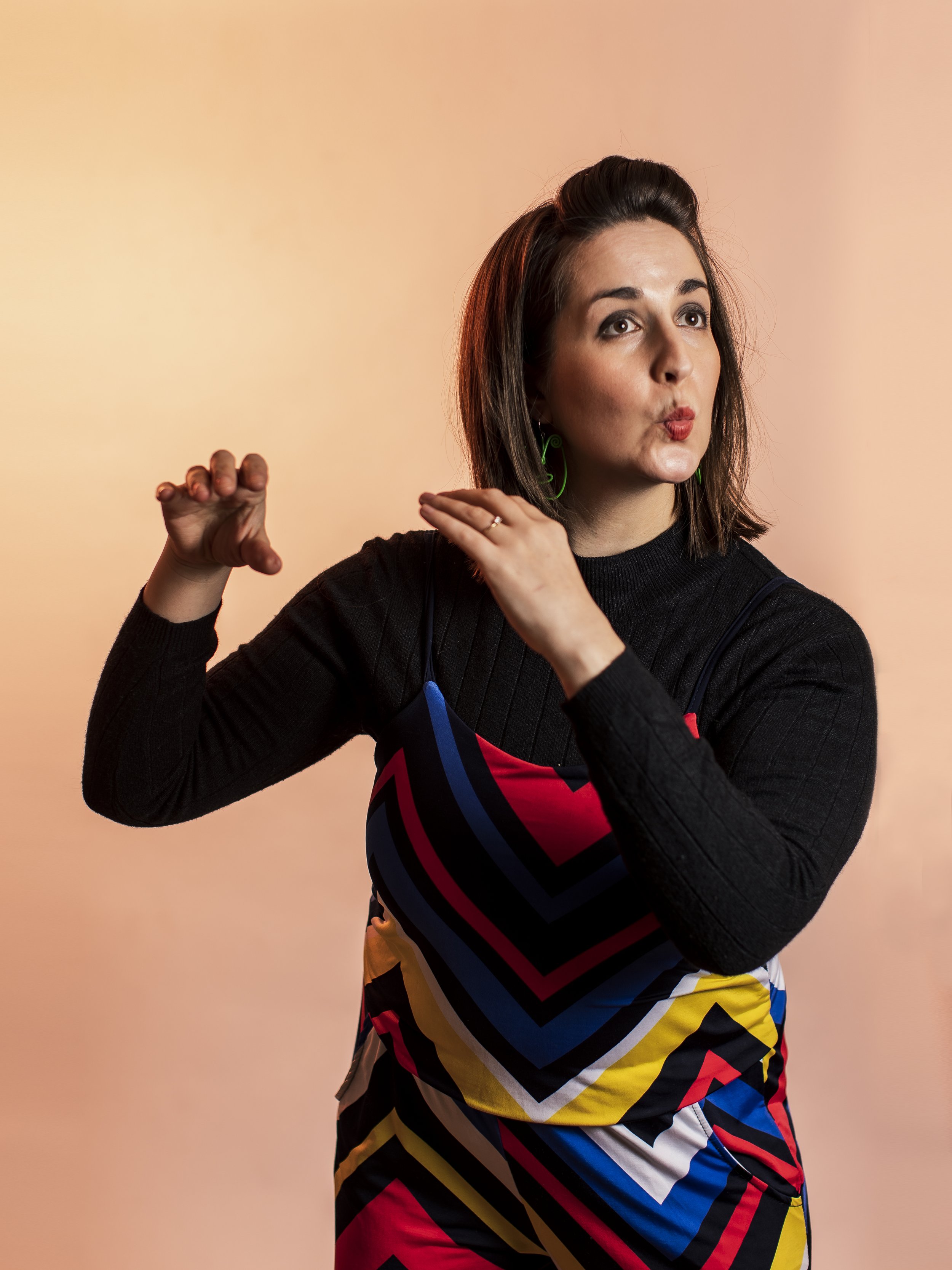 Woman with shoulder-length brown hair, wearing a colorful dress with geometric patterns and a black long-sleeve top, making a playful gesture and pursing her lips.