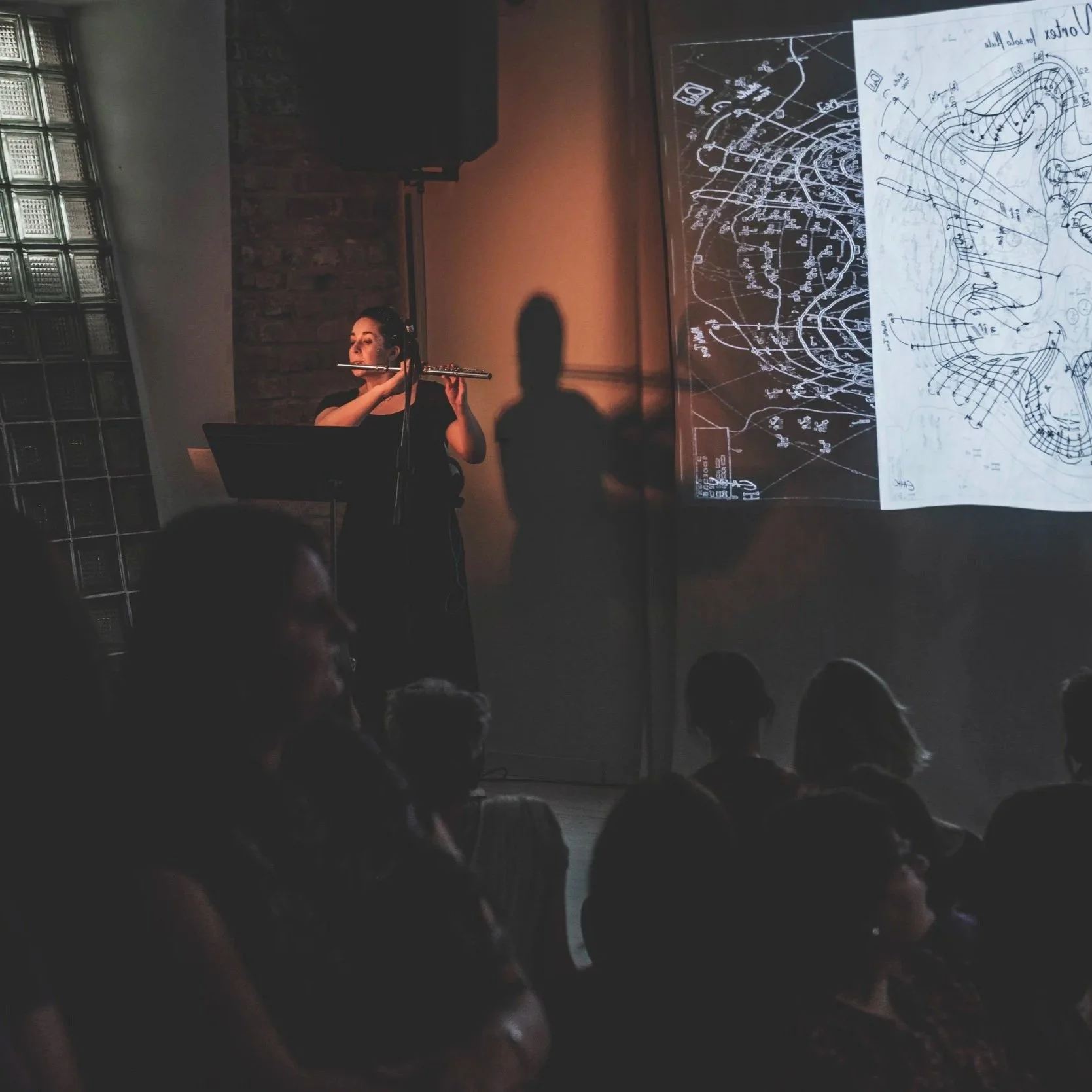 A woman playing flute on stage during a presentation with maps projected on a screen behind her, audience seated in foreground.