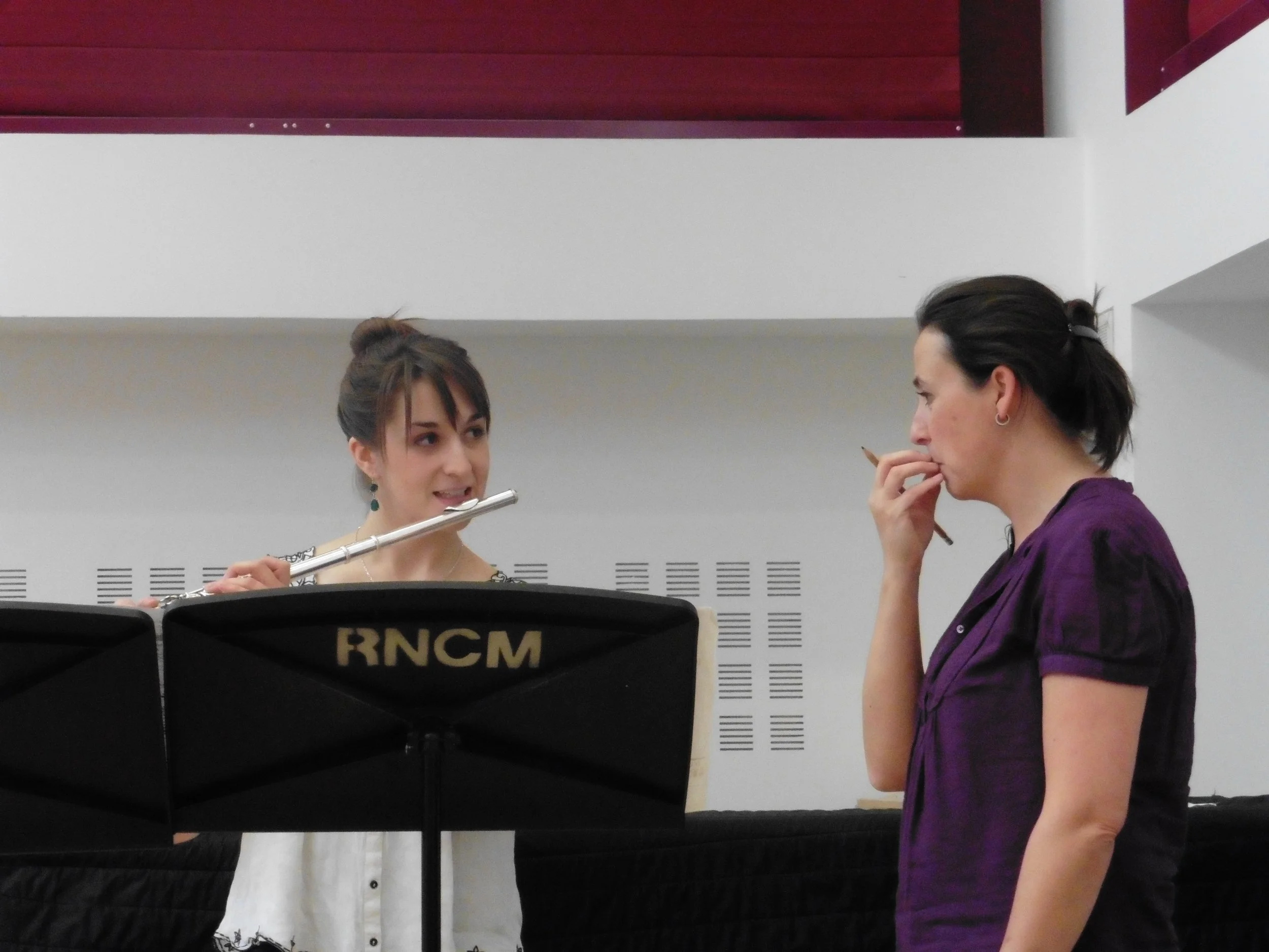 Two women having a discussion in a music rehearsal or lesson, one holding a flute, with music stand labeled RNCM in front of them.