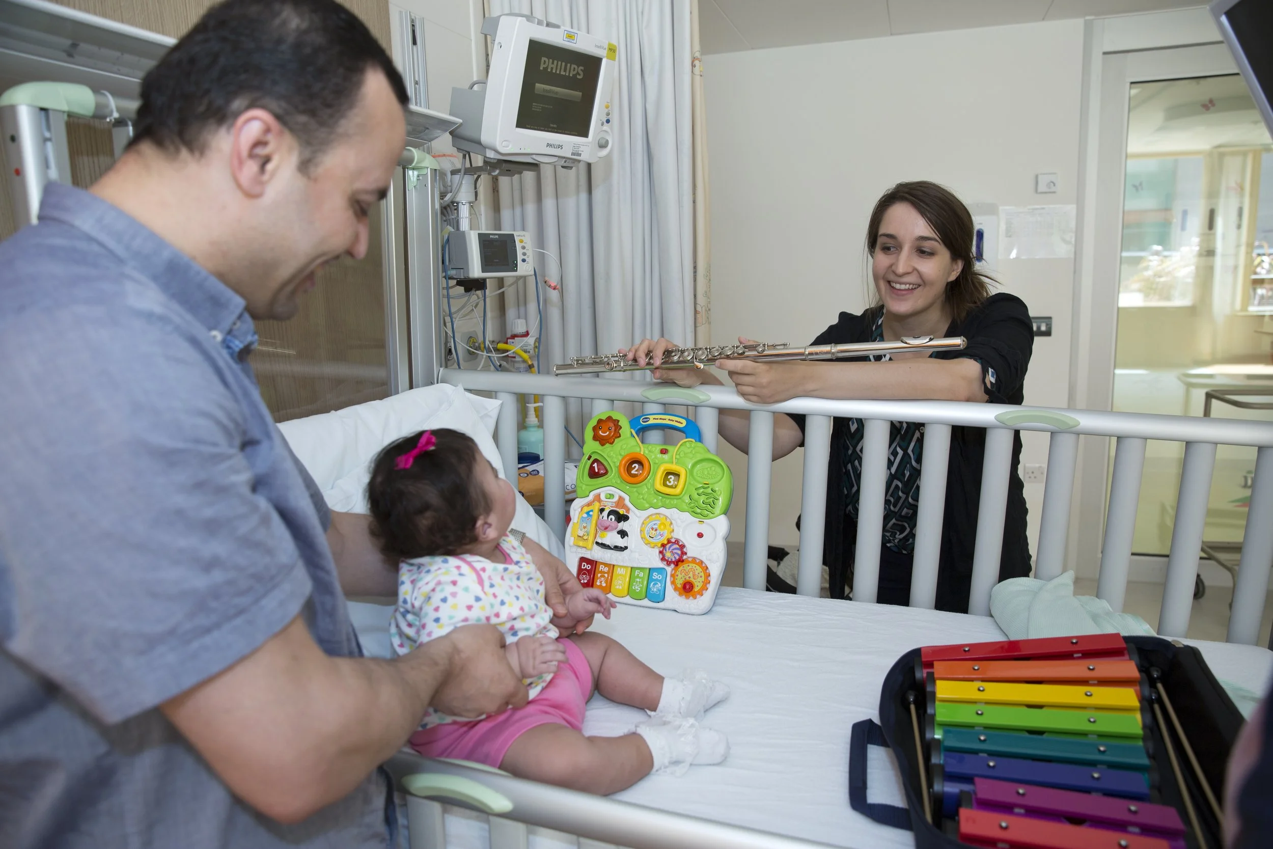 A young girl in a hospital bed with a man, possibly her father, holding her hands, and a woman, likely a music therapist, playing a flute across the bed. Colorful toys and a xylophone are also on the bed.
