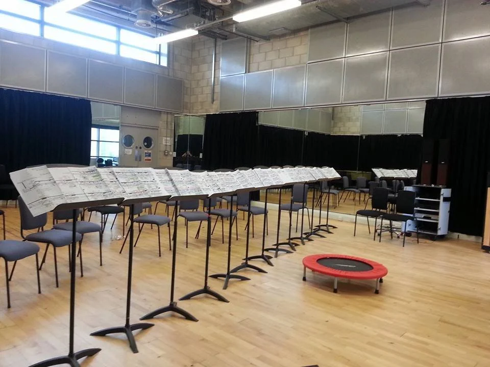 Large room with a row of music stands, chairs, and a stack of sheet music or newspapers, black curtains, a small trampoline, and a corner with mirrors and equipment.
