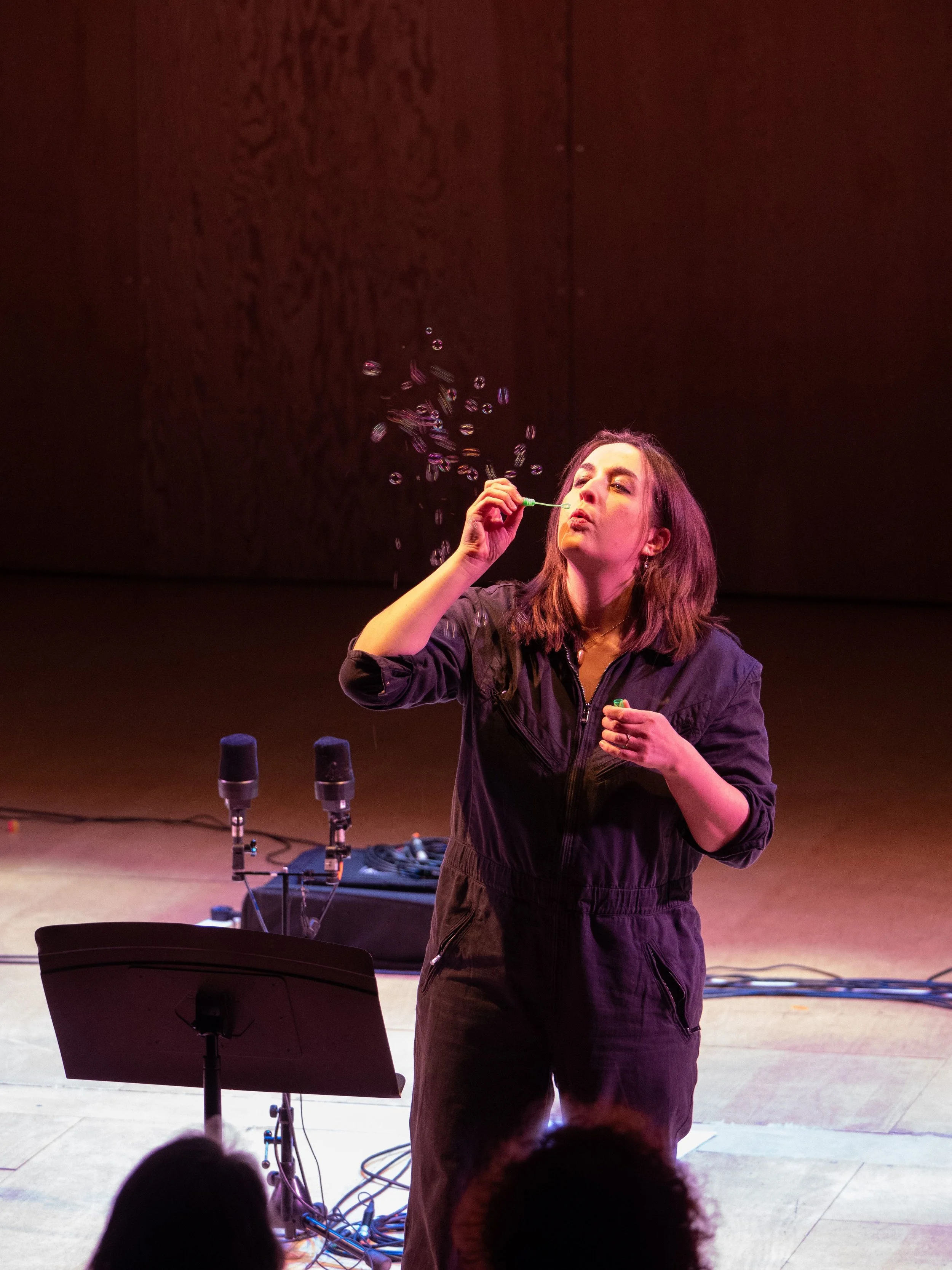 A woman in a black jumpsuit blowing bubbles on a stage with microphones and music equipment in front of her.