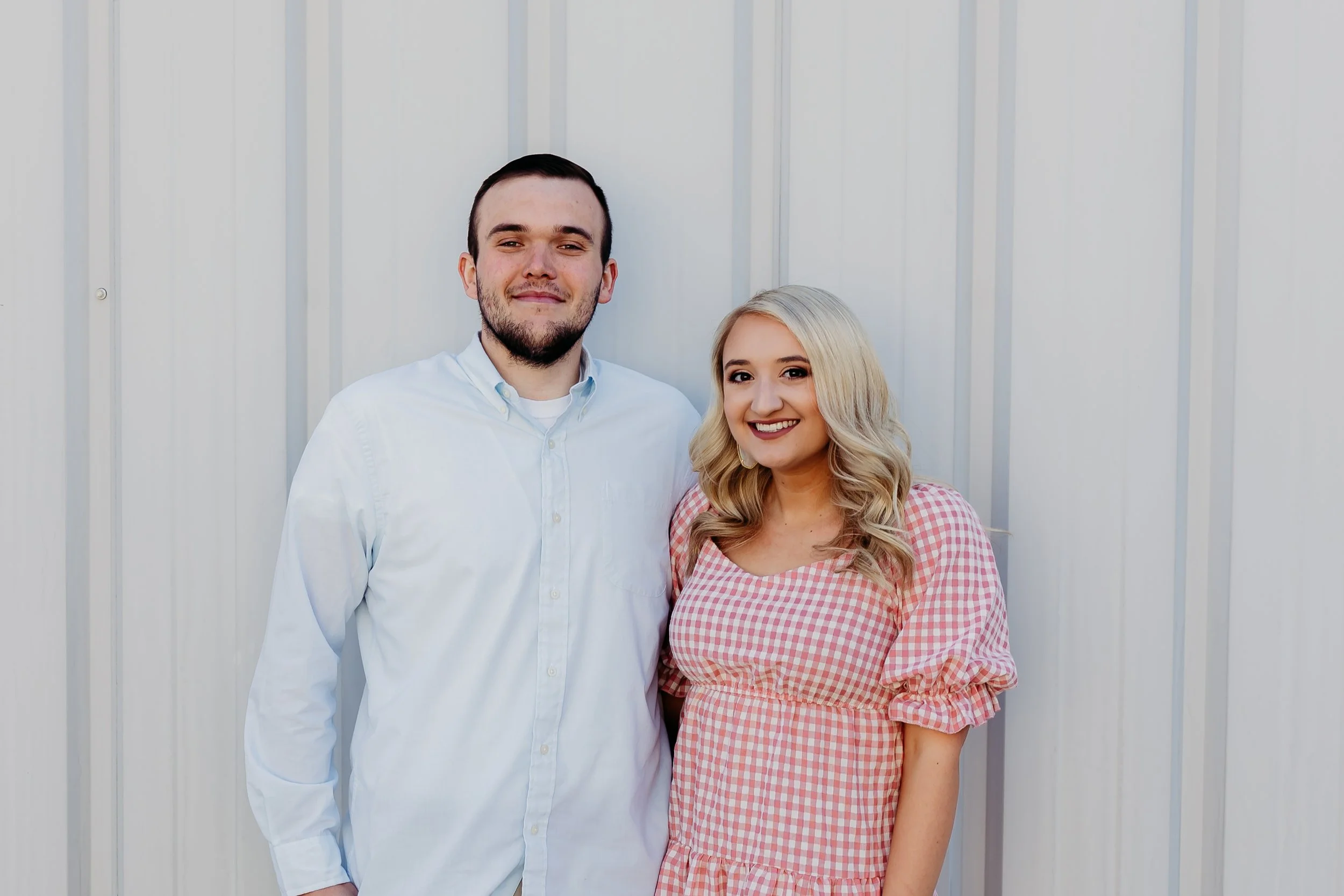 A young man and woman standing side by side outdoors, smiling, with a light-colored metal wall in the background.