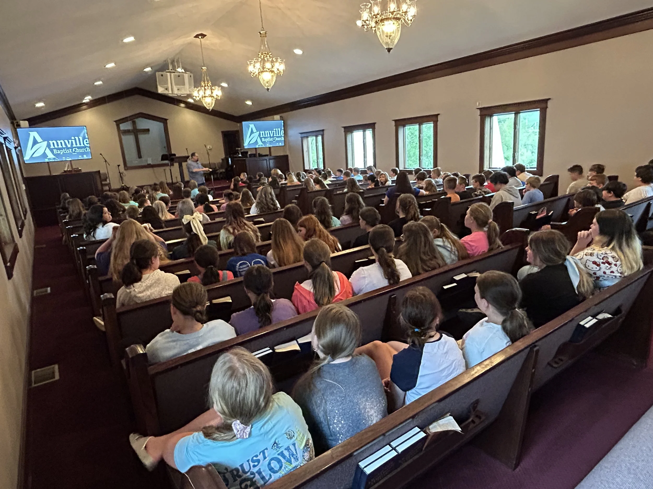 A crowded church congregation seated in pews during a service or event, with a speaker at the front and two screens displaying the Annville Baptist Church logo.