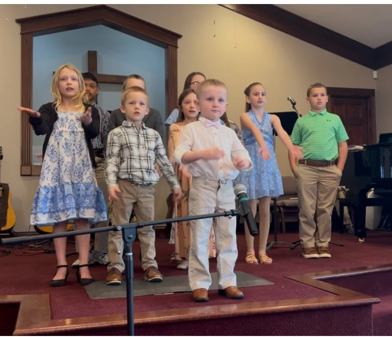 A group of children singing or performing on a small stage inside a church or community hall, some with microphones, dressed in semi-formal attire.