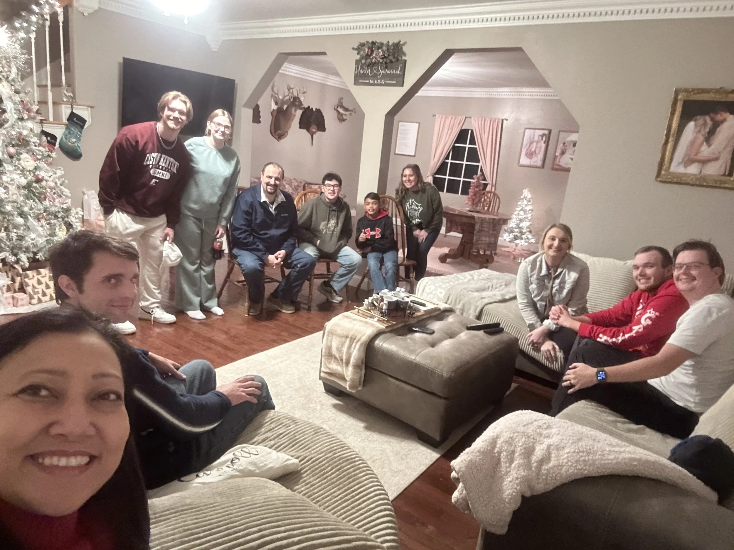 A group of people gathered in a living room decorated for Christmas, with a Christmas tree, stockings, and festive decorations.