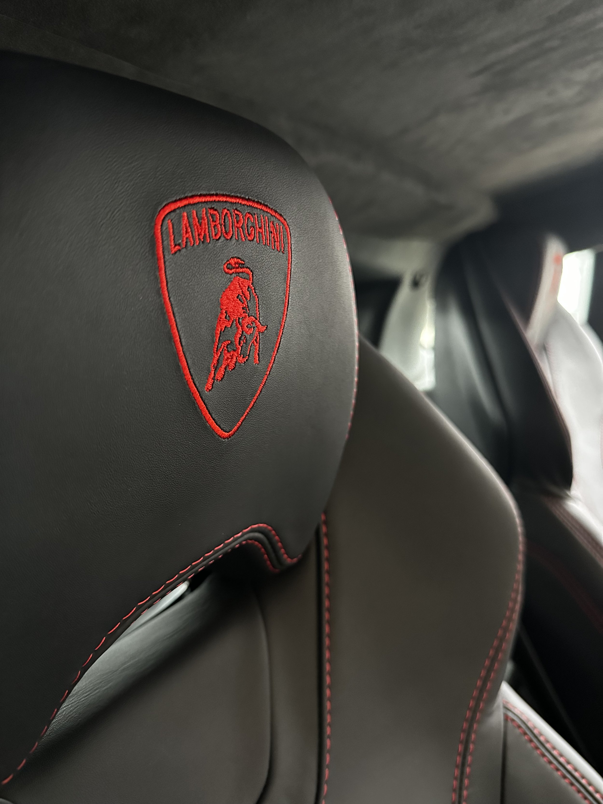 Close-up of black leather car seat headrest with red embroidered Lamborghini logo and red stitching.