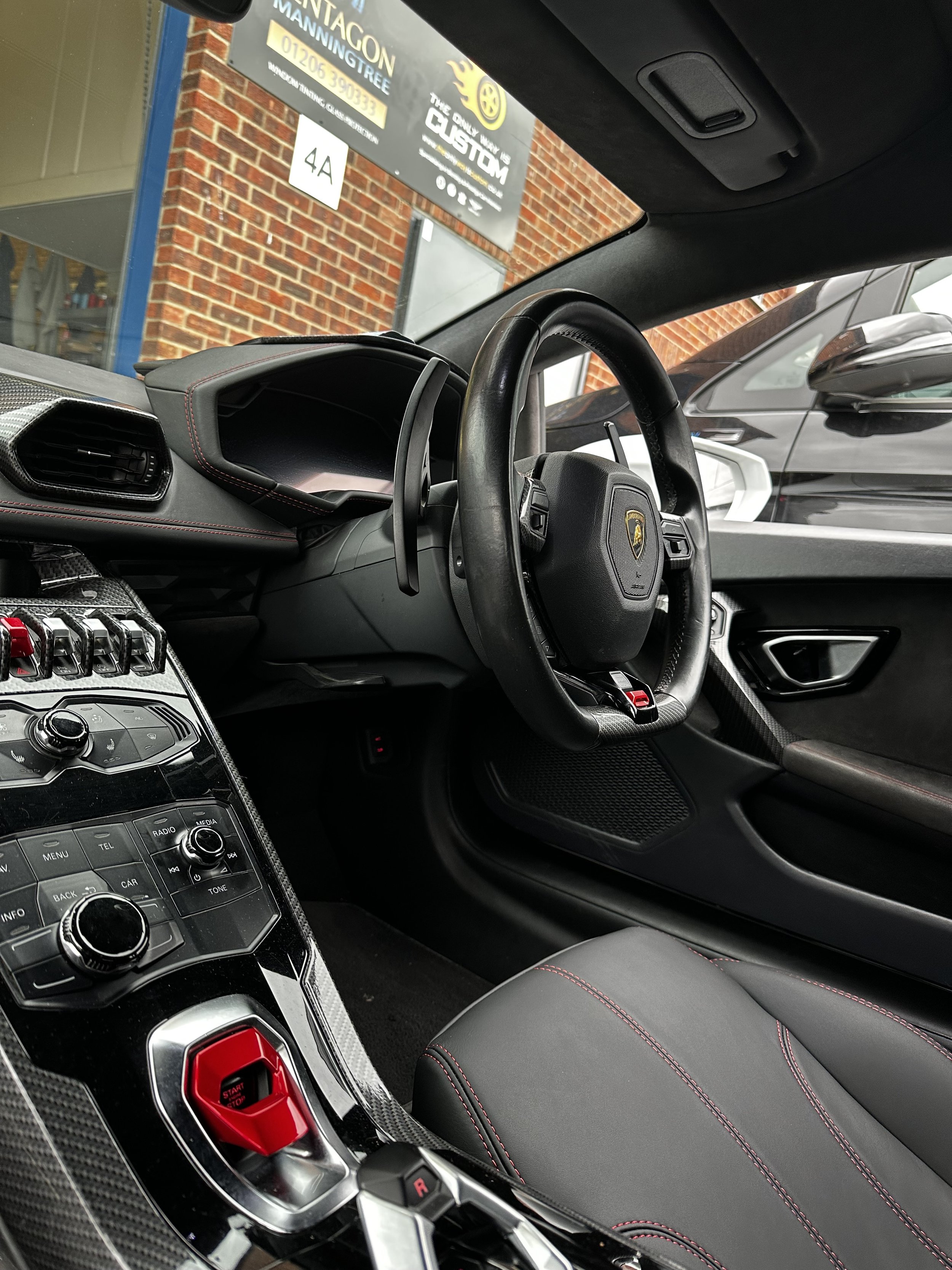 Interior of a luxury Lamborghini sports car showing a black leather steering wheel, dashboard, carbon fiber center console with red accents, and black leather seat with red stitching.