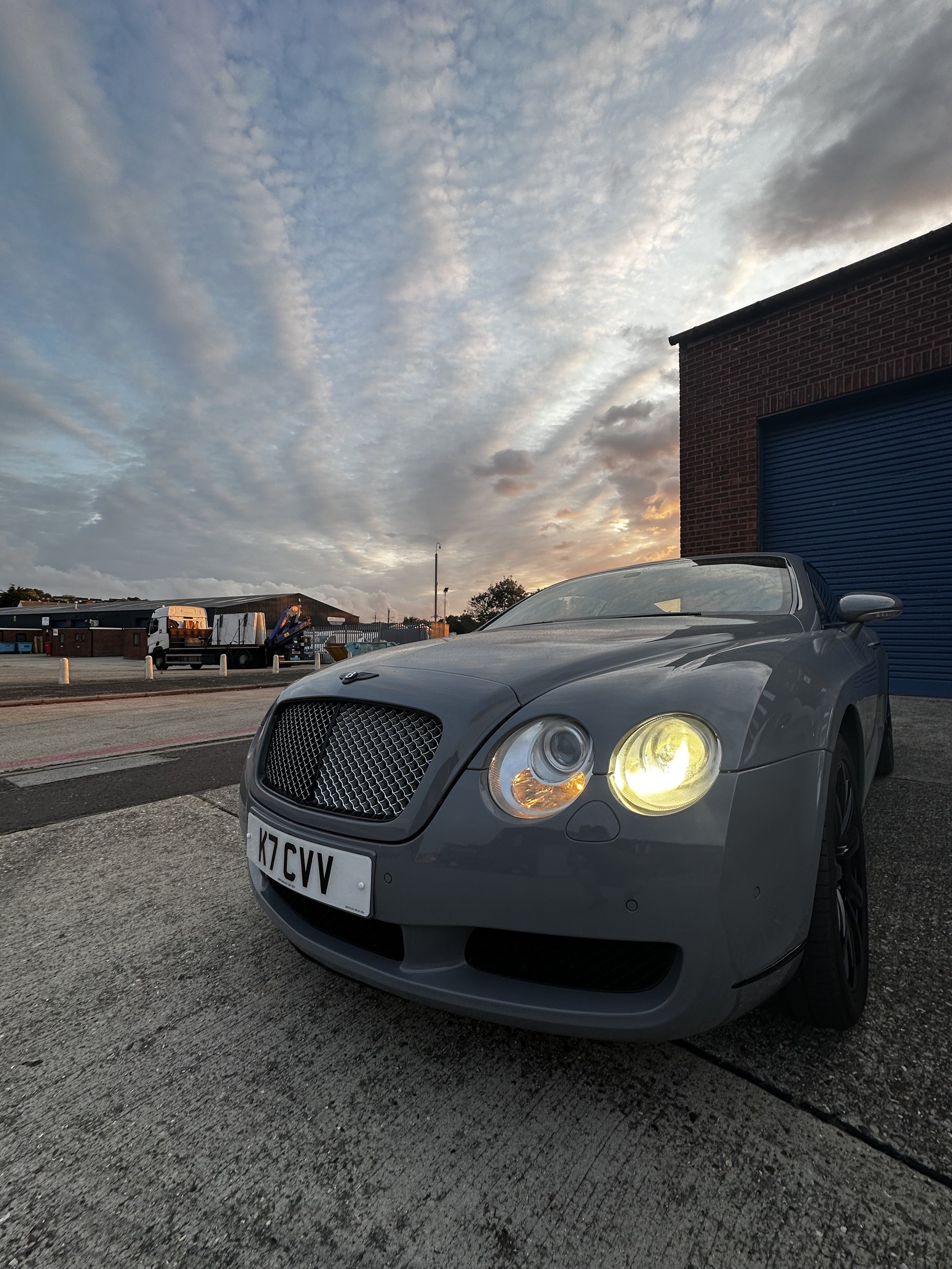 A silver Bentley Continental car parked on a concrete surface near a brick building with a blue garage door, during sunset with a partly cloudy sky in the background.