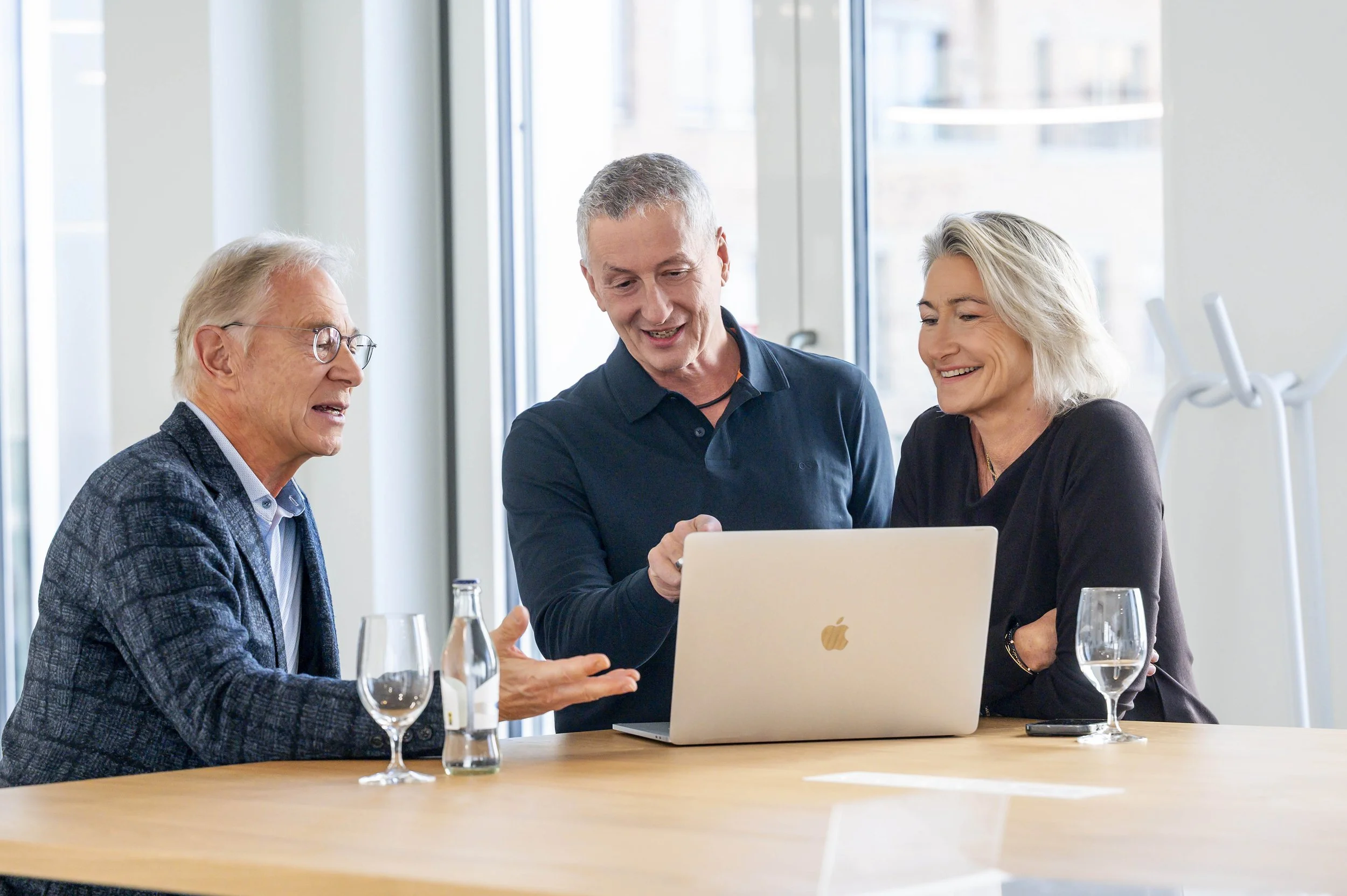 Drei Menschen sitzen an einem Tisch, schauen auf einen Laptop und tauschen sich aus in einem hellen Raum.