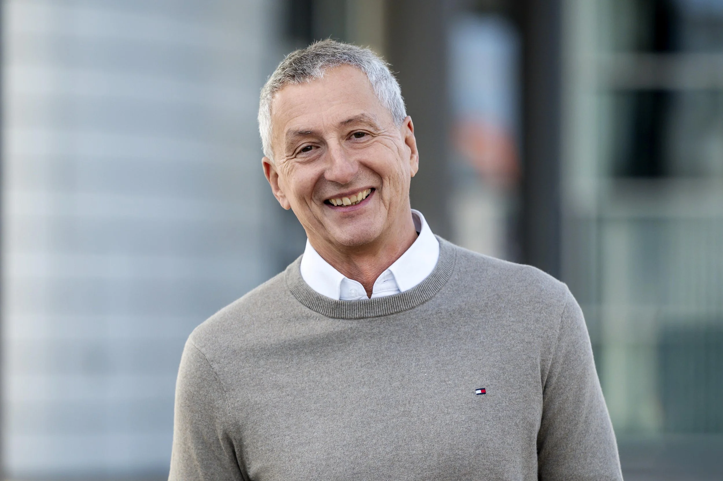 Ein lächelnder älterer Mann trägt einen weißen Hemd und grauen Pullover mit einem kleinen Tommy Hilfiger Logo, steht vor einem modernen Gebäudekomplex, Außenaufnahme.