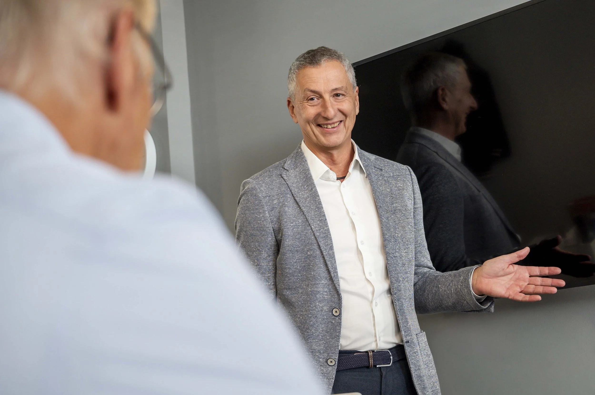 Ein Mann in einem grauen Sakko und weißem Hemd lächelt und gestikuliert bei einer Präsentation, während ein anderer Mann im Vordergrund sitzt.