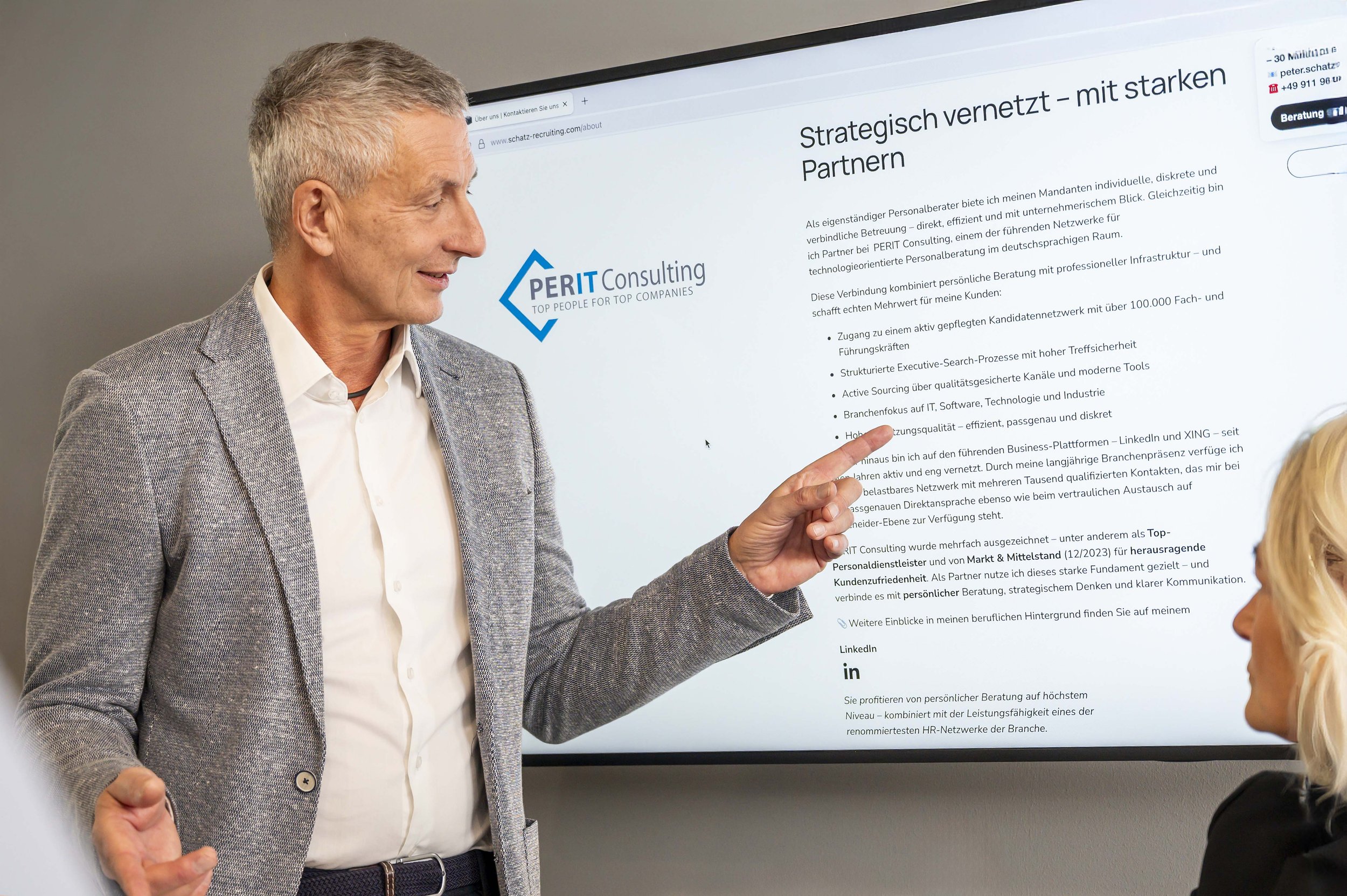 Ein Mann in einem grauen Anzug erklärt etwas auf einer Präsentation auf einem Bildschirm, während eine Frau zuhört. Der Bildschirm zeigt das Logo und Text von PERIT Consulting.