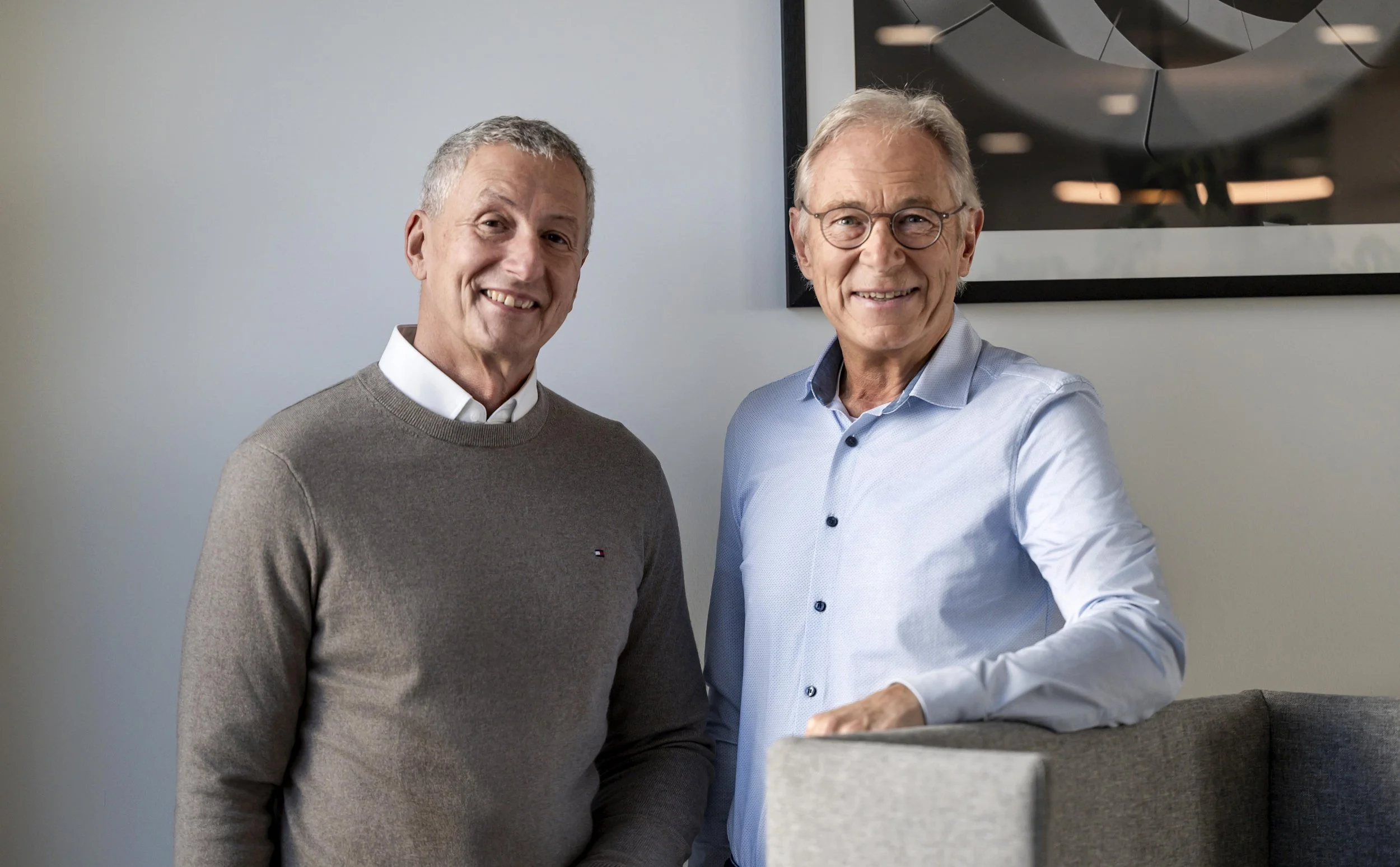 Zwei Männer stehen nebeneinander, lächeln und posieren vor einer Wand mit einem schwarzen Rahmen. Einer trägt einen grauen Pullover und der andere ein hellblaues Hemd.