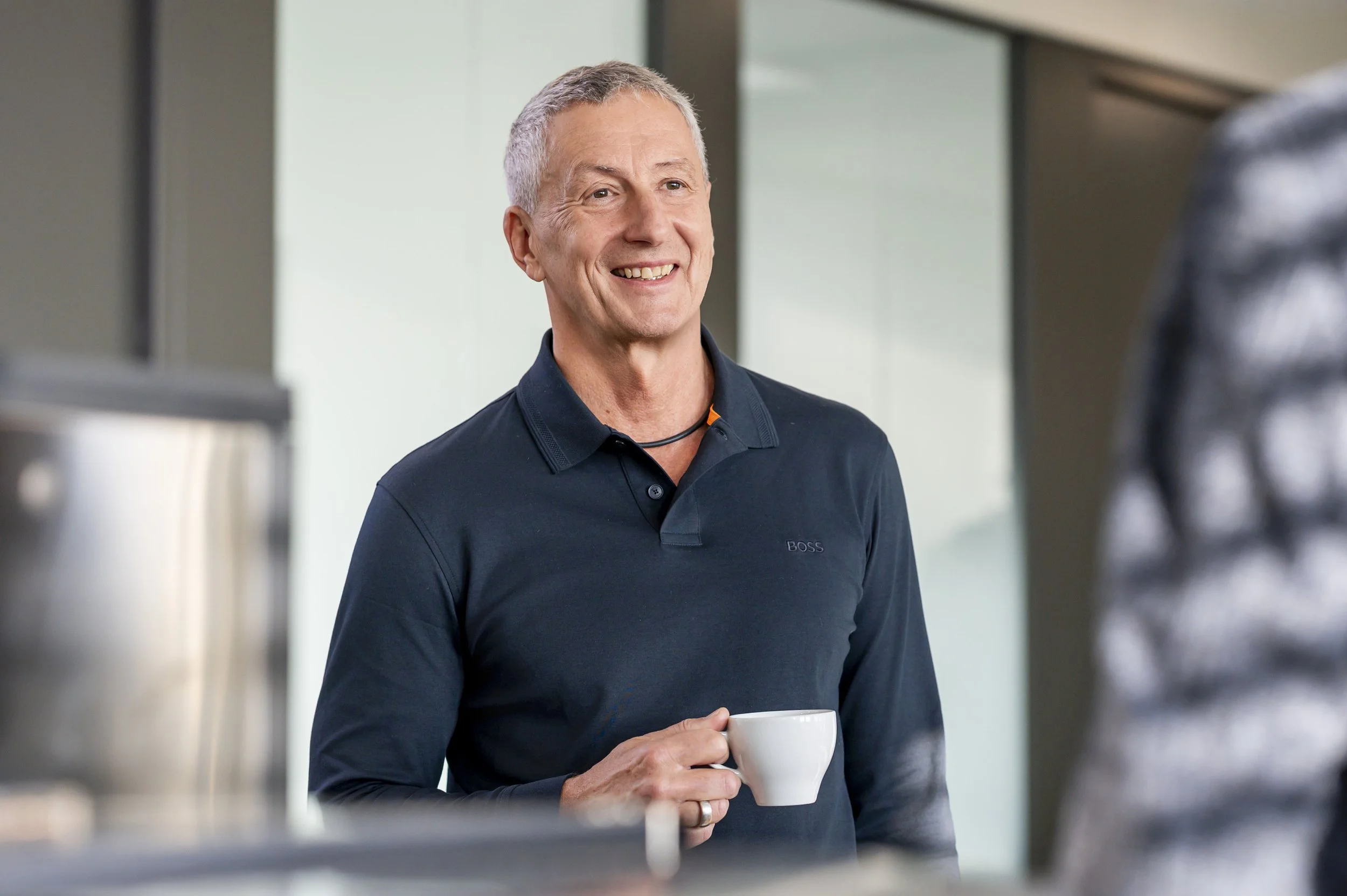 Peter Schatz, lächelnd, hält eine weiße Kaffeetasse in der Hand, steht in einem modernen Büro oder Café.