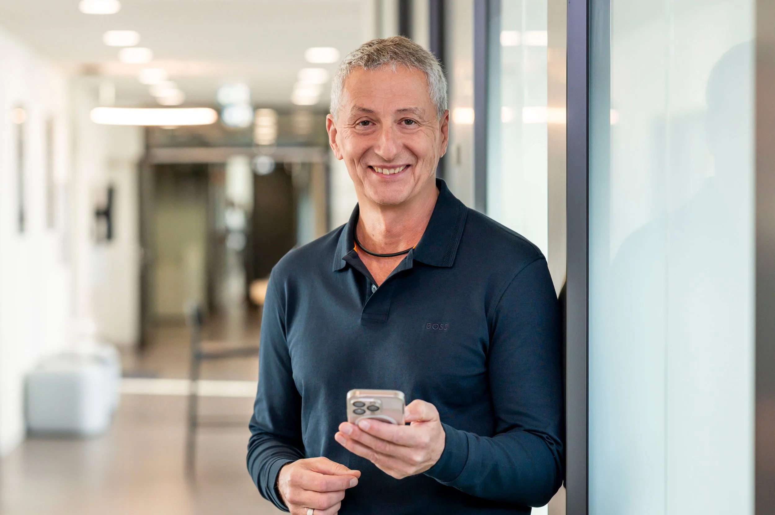 Ein Mann lächelt, während er ein Smartphone in der Hand hält, in einem modernen Büroflur.
