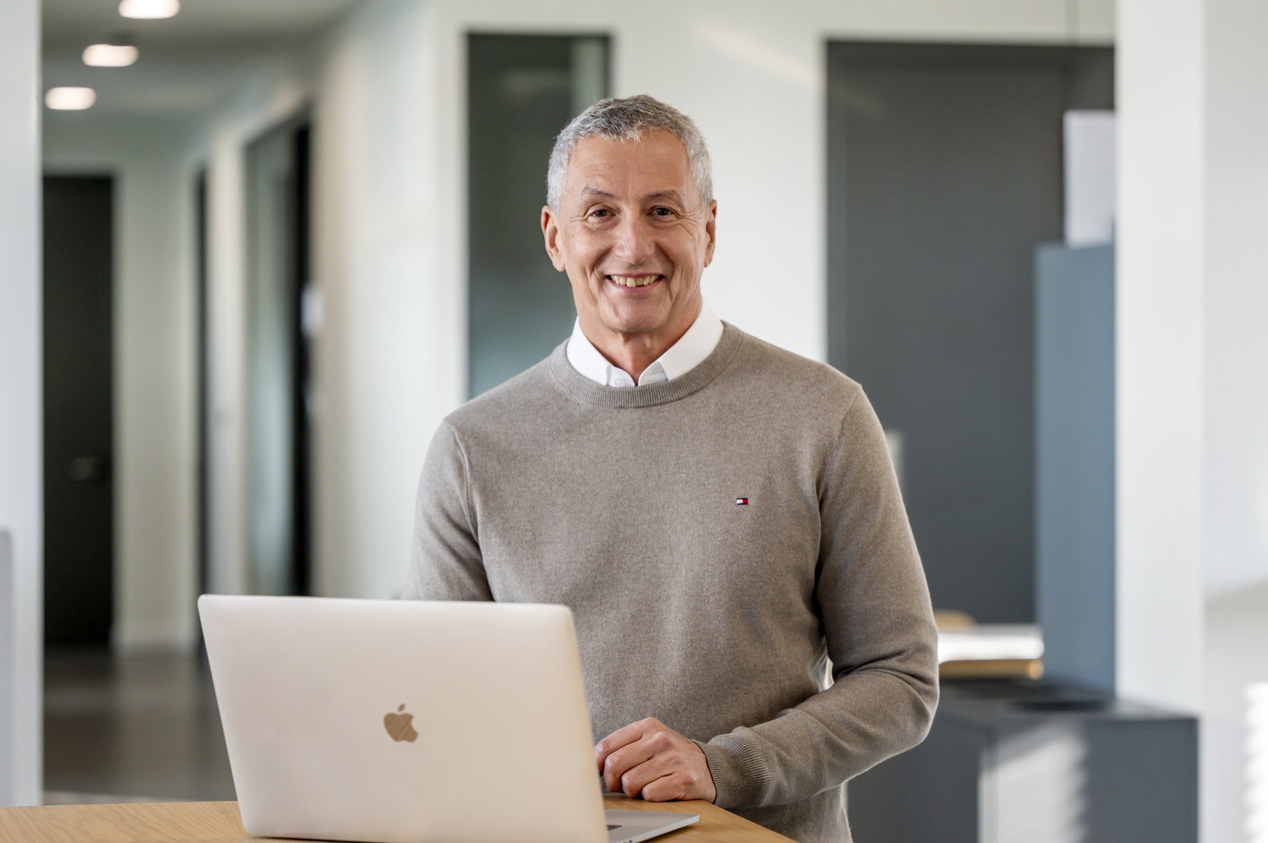 Ein lächelnder Mann steht hinter einem weißen Apple-Laptop auf einem Holzschreibtisch in einem modernen Büro, trägt einen beigen Pullover mit Markenlogo und eine weiße Hemdkragen, im Hintergrund sind weiße Wände und dunkle Türöffnungen sichtbar.