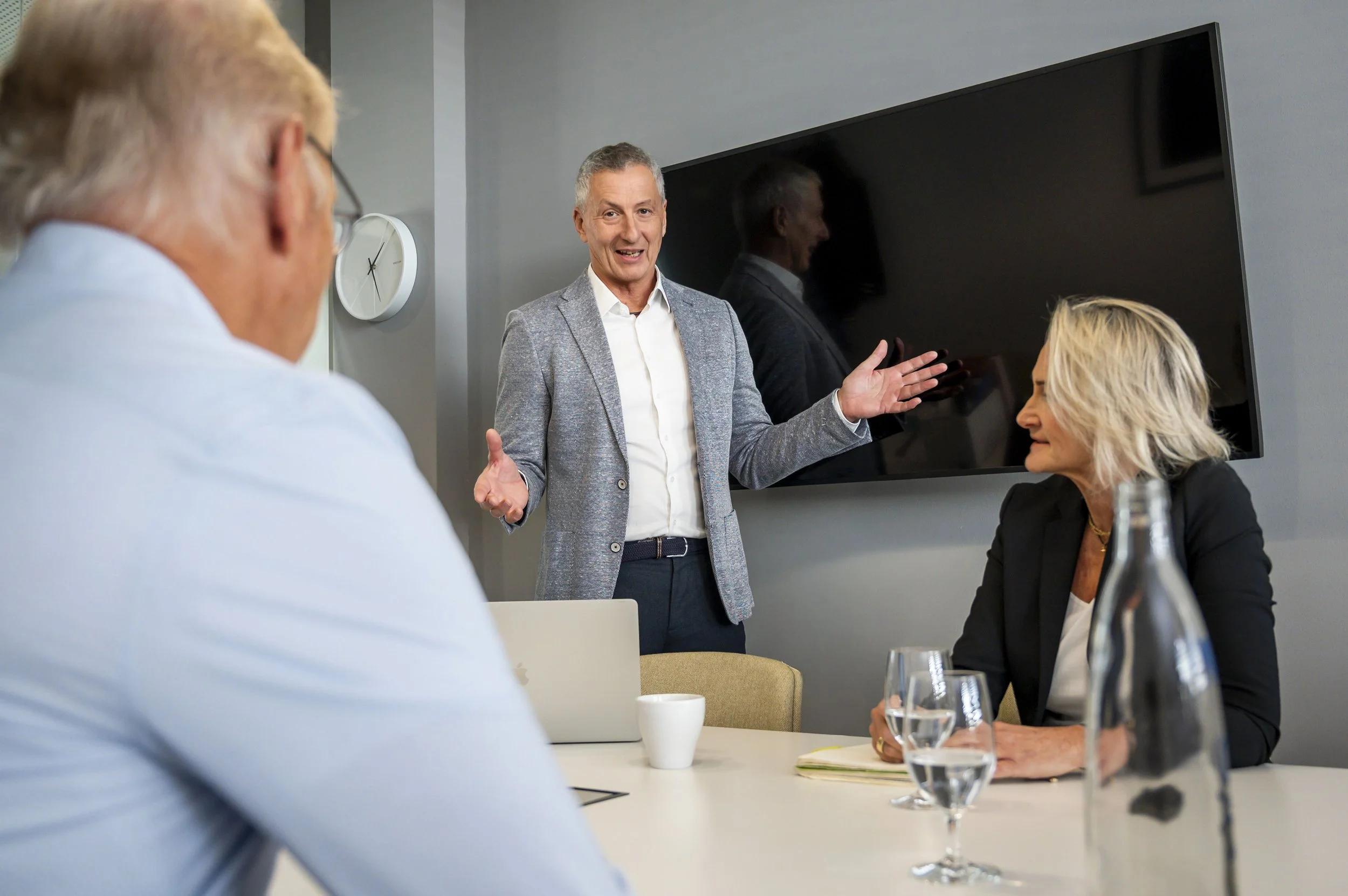 Ein Mann steht während einer Besprechung vor einem großen Bildschirm und spricht zu zwei anderen Personen, während sie an einem Konferenztisch sitzen, mit Wasser und Notizen.