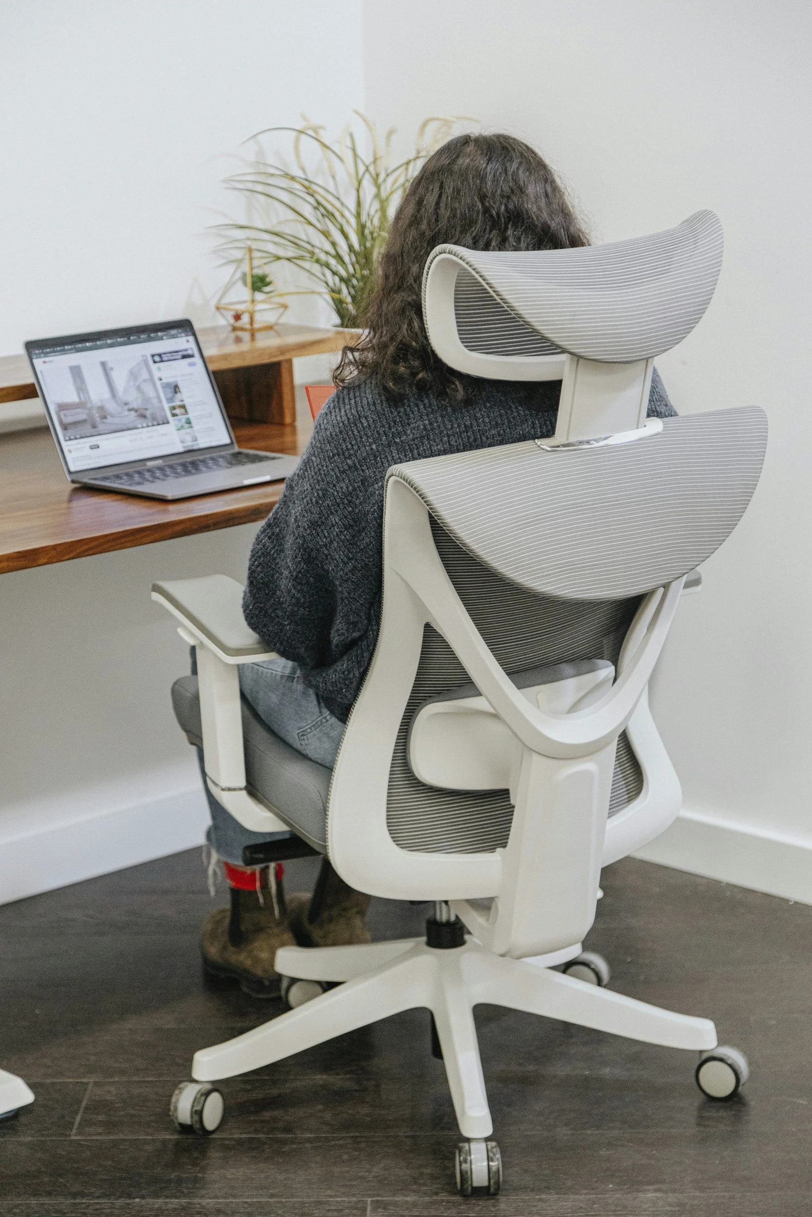 A person with curly dark hair sitting on an ergonomic white office chair at a wooden desk, working on a laptop in a modern home office space.