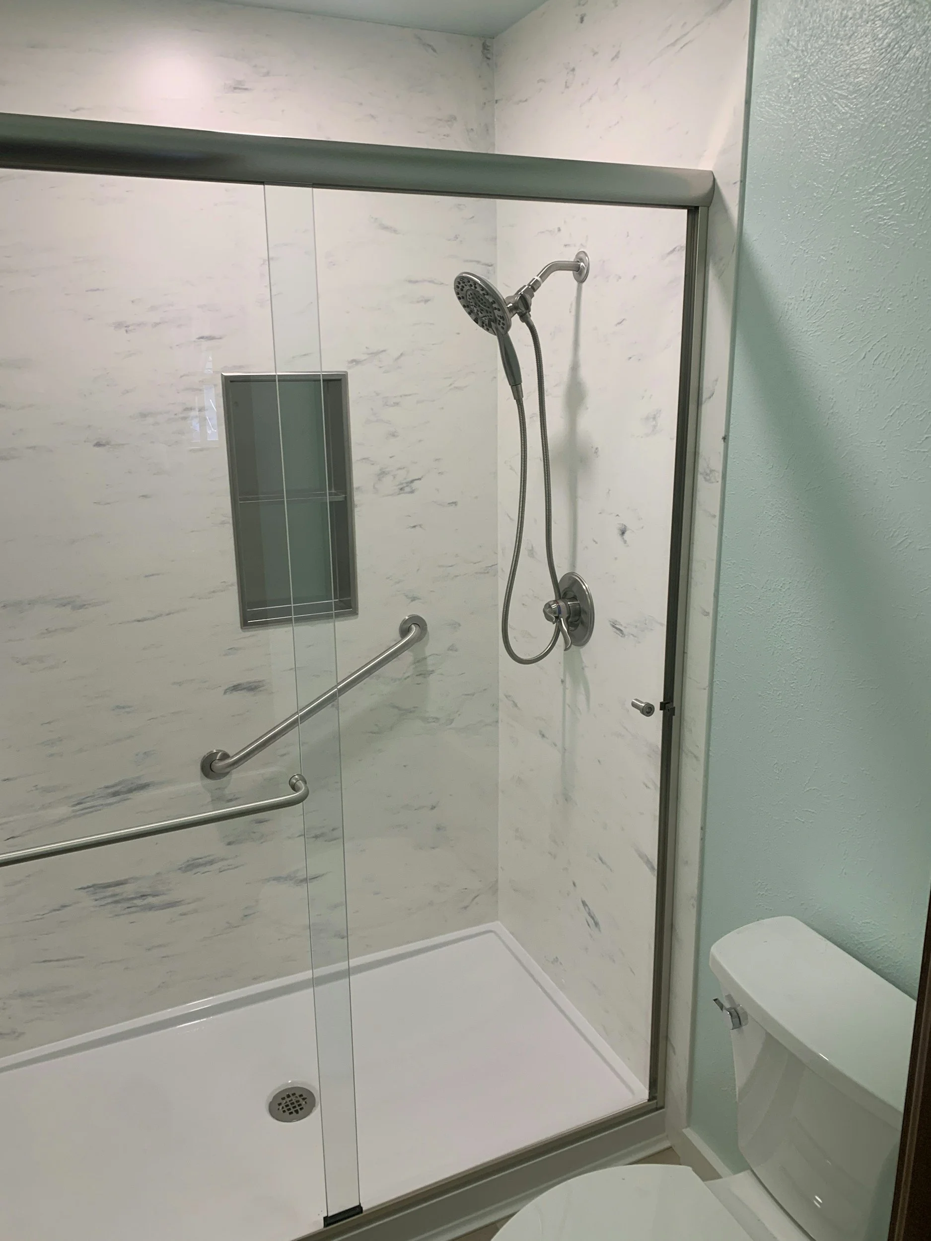 A walk-in shower with a sliding glass door, marble walls, a showerhead, and a grab bar, next to a toilet.