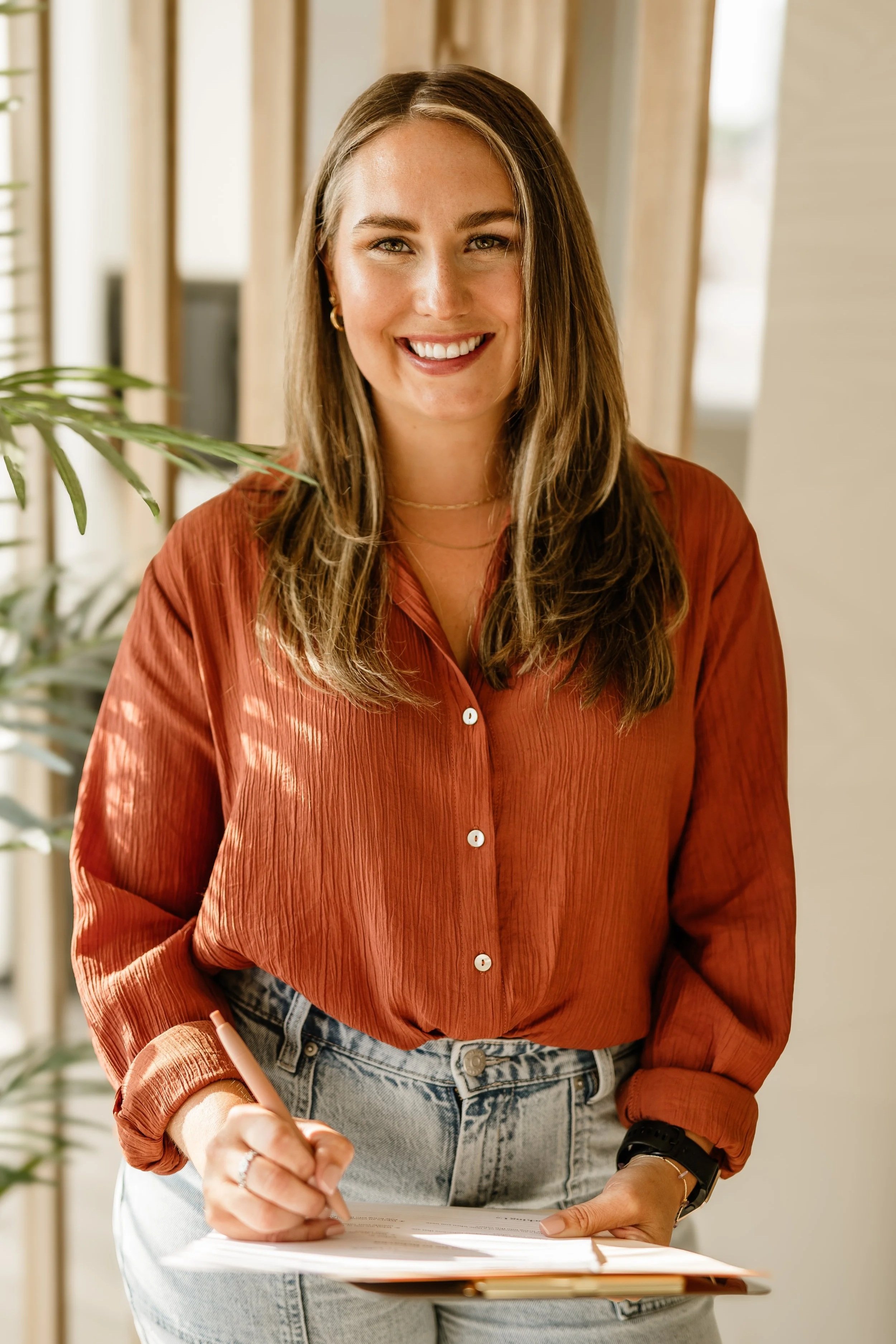 A woman with long, wavy brown hair wearing a rust-colored blouse and light blue jeans, smiling while holding a clipboard and pen inside a well-lit room with beige curtains and a plant.
