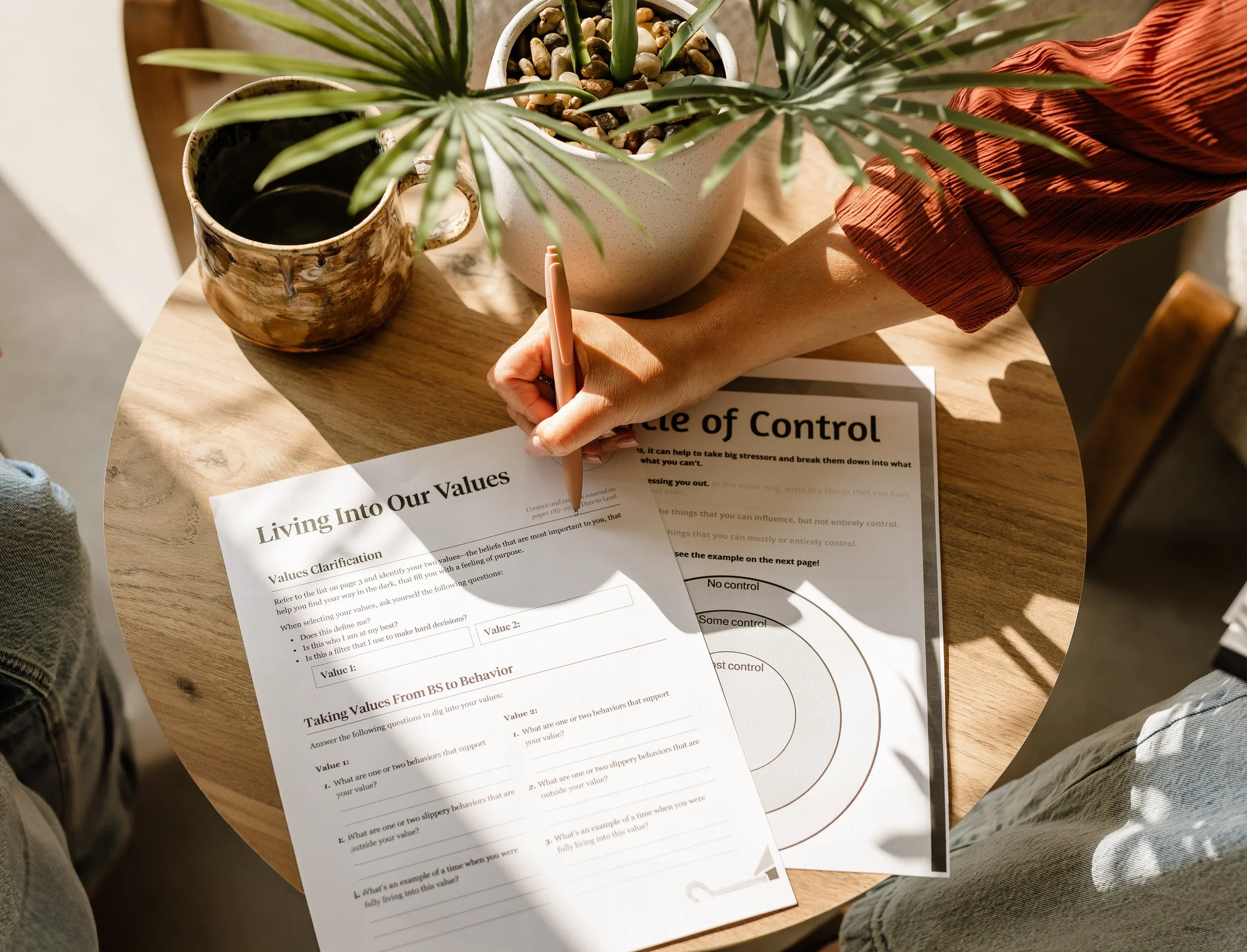 A person with a reddish sleeve is writing on a sheet titled "Living Into Our Values" on a wooden table, surrounded by potted plants, sunlight creating shadows.