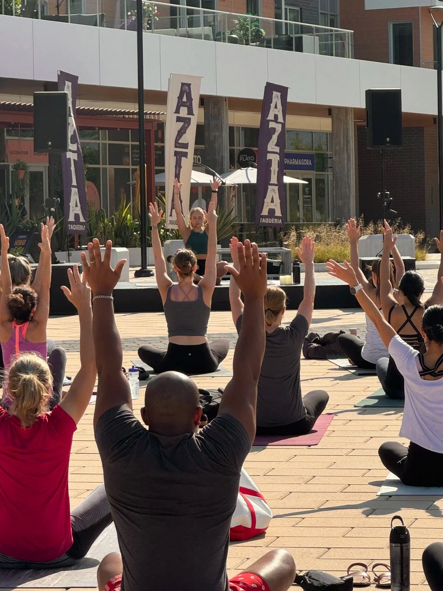 Des personnes participant à un cours de yoga en plein air, les bras levés, face à une scène où une femme dirige la séance, devant des banderoles sur lesquelles on peut lire "AZTIA Taquería".