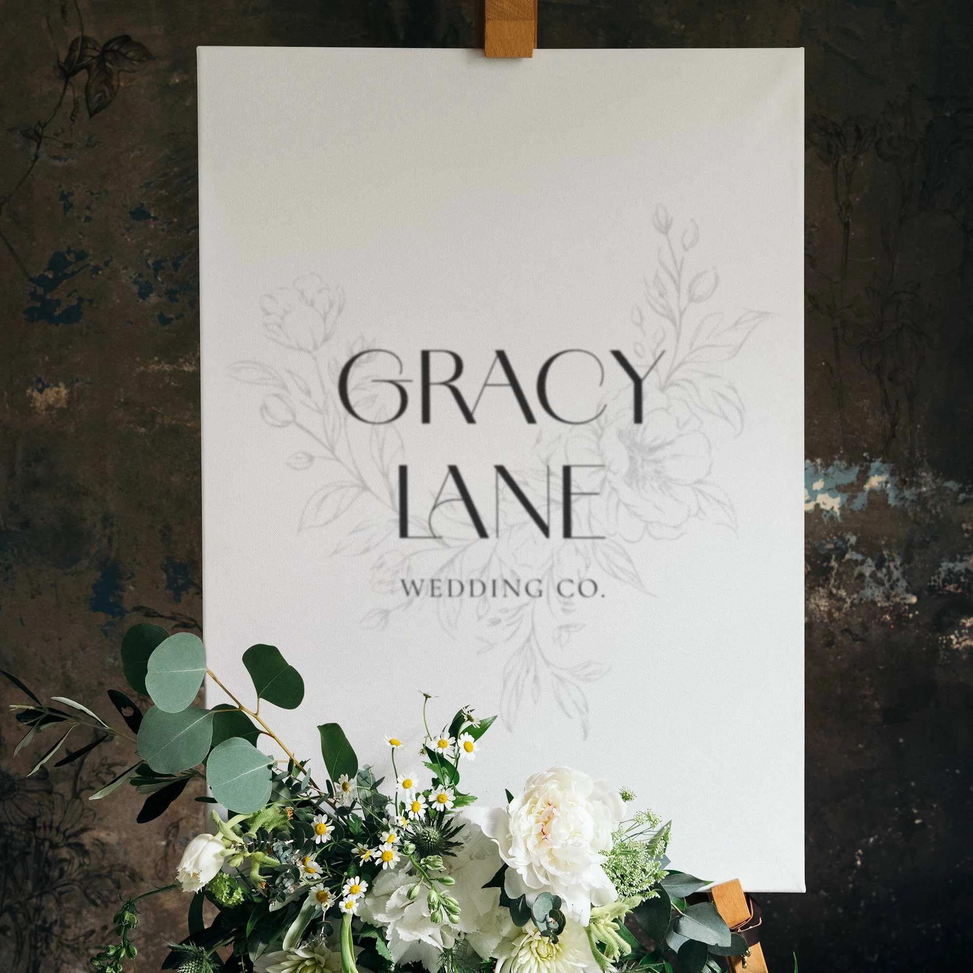 White sign with black text reading 'Gracy Lane Wedding Co.' behind a floral arrangement of white flowers and greenery, on a dark textured background.