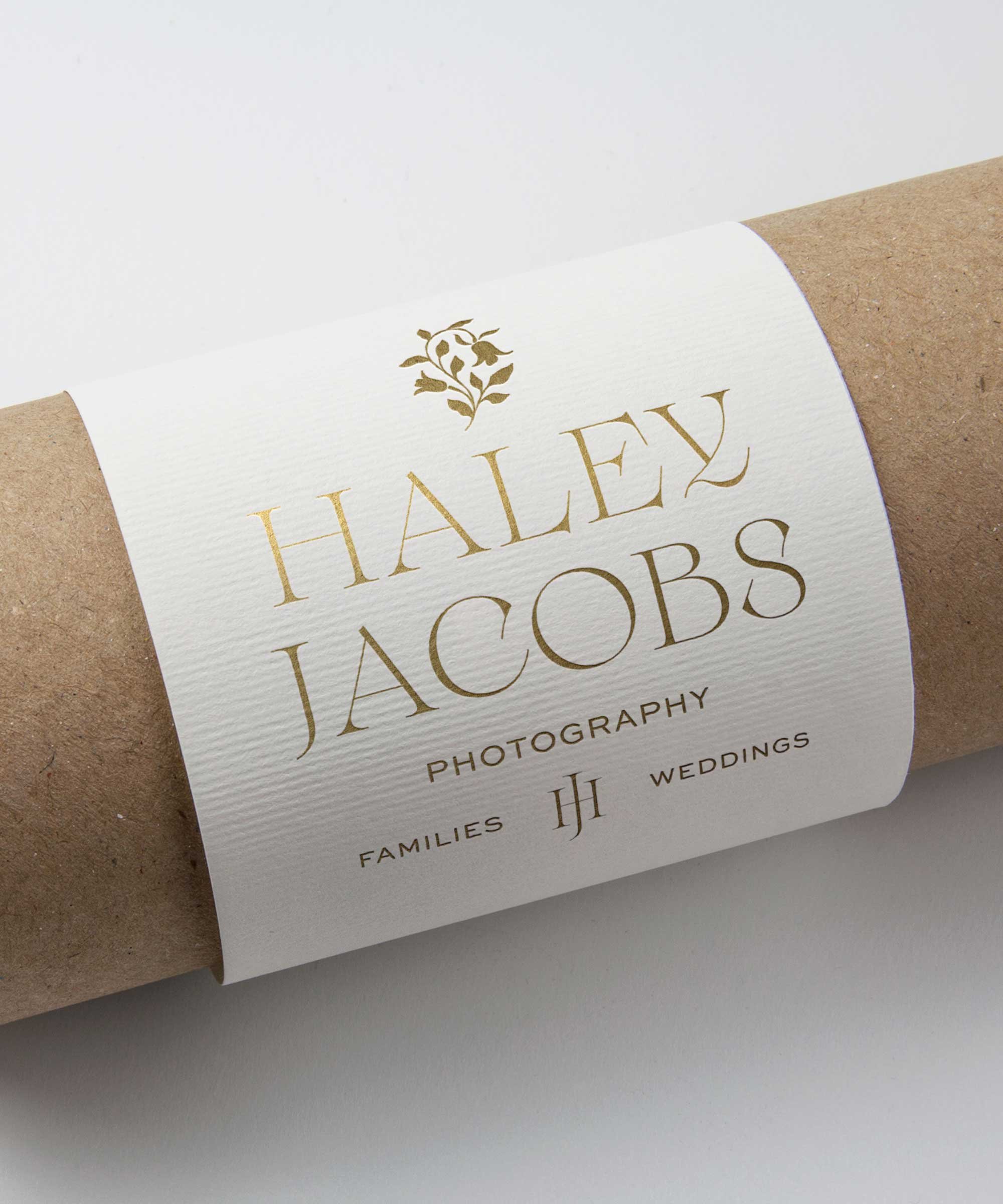 Roll of paper with a white label featuring gold lettering that says Gothic style 'HALEY JACOBS' and smaller text below reading 'PHOTOGRAPHY', 'FAMILY', 'WEDDINGS', with a decorative floral icon above and initials 'HJ' in between.