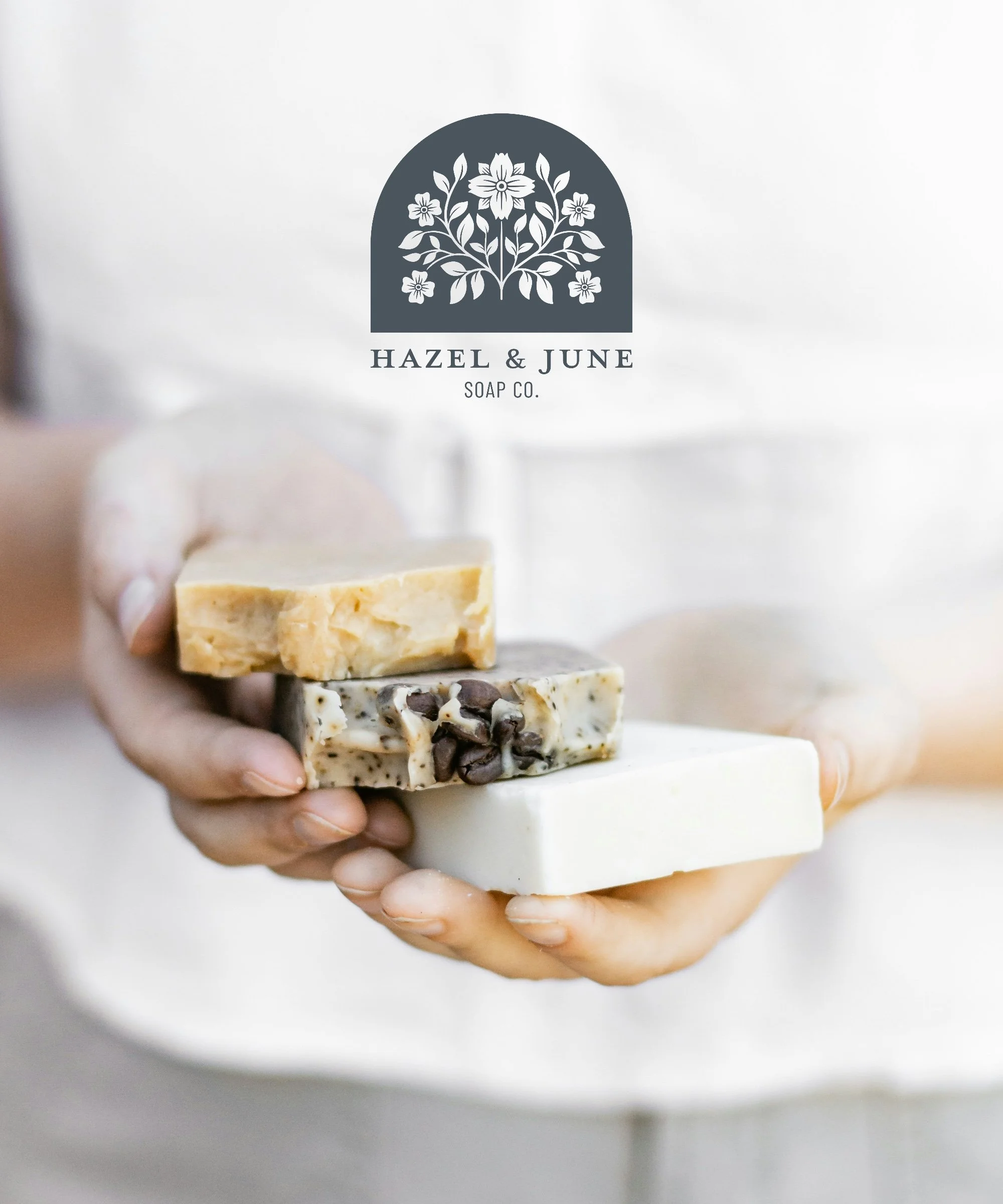 A person holding four different handmade soap bars in front of a logo for Hazel & June Soap Co. The soaps are in various shades and textures, including one with chocolate chips.