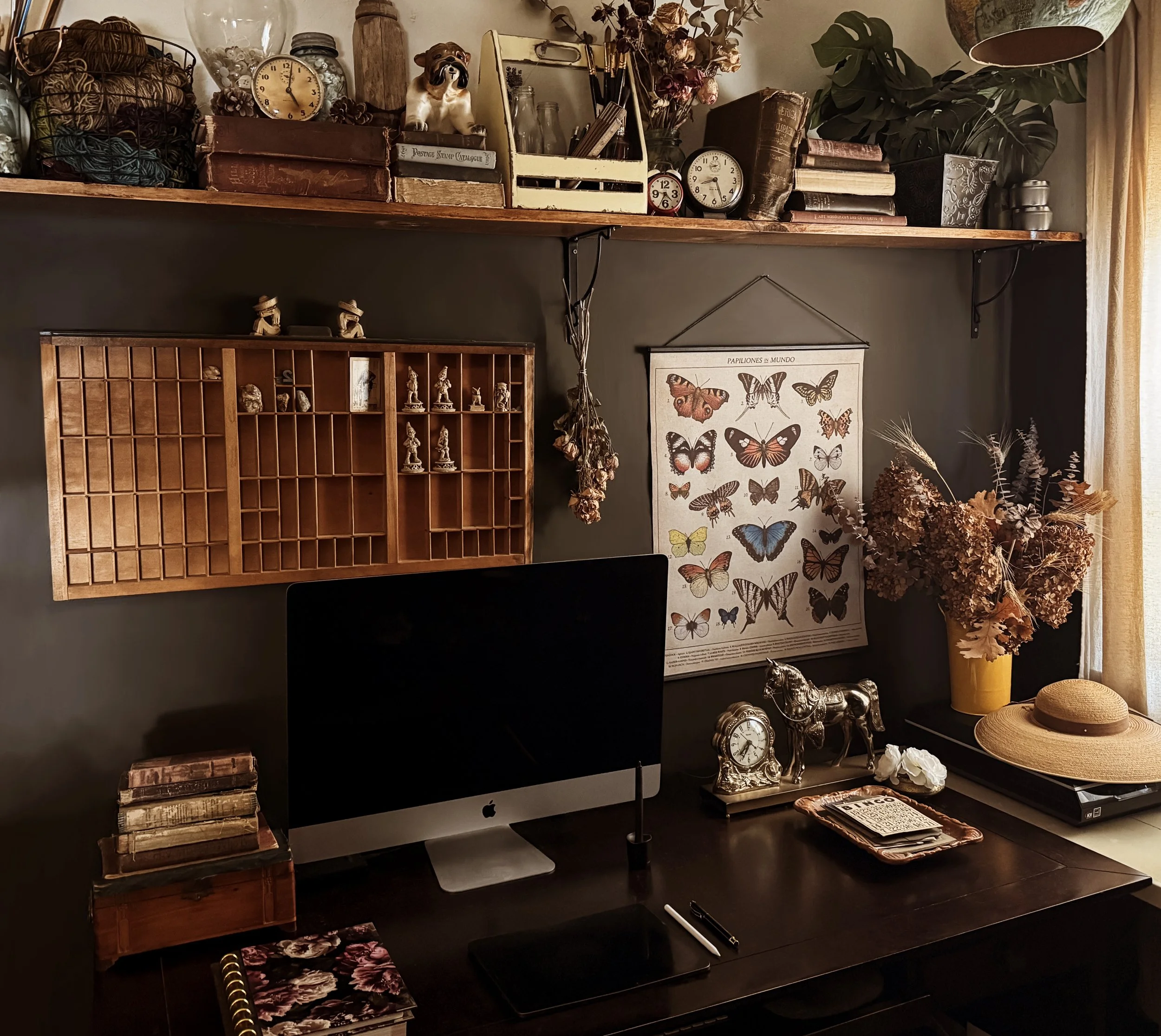Home office desk with a computer, decorative items, dried flowers, a hat, and butterfly poster on the wall.