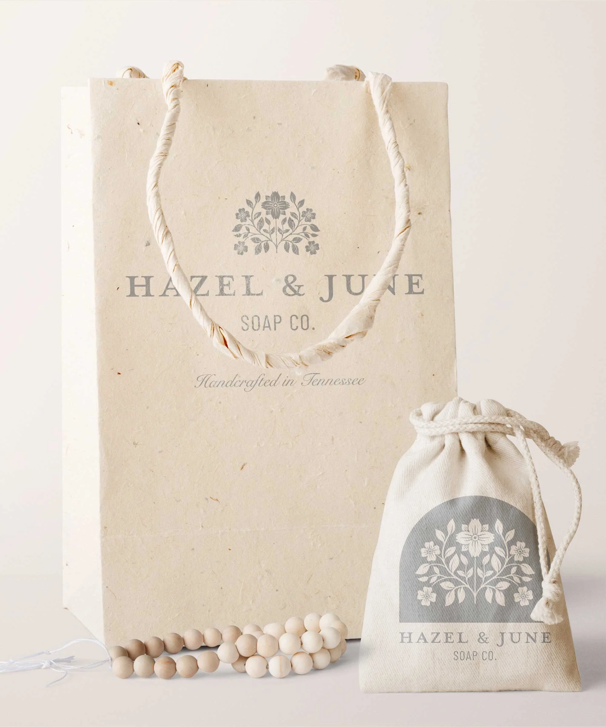 A large paper shopping bag and a smaller fabric gift pouch, both with the Hazel & June Soap Co. logo and a floral design, along with a beaded bracelet in front.