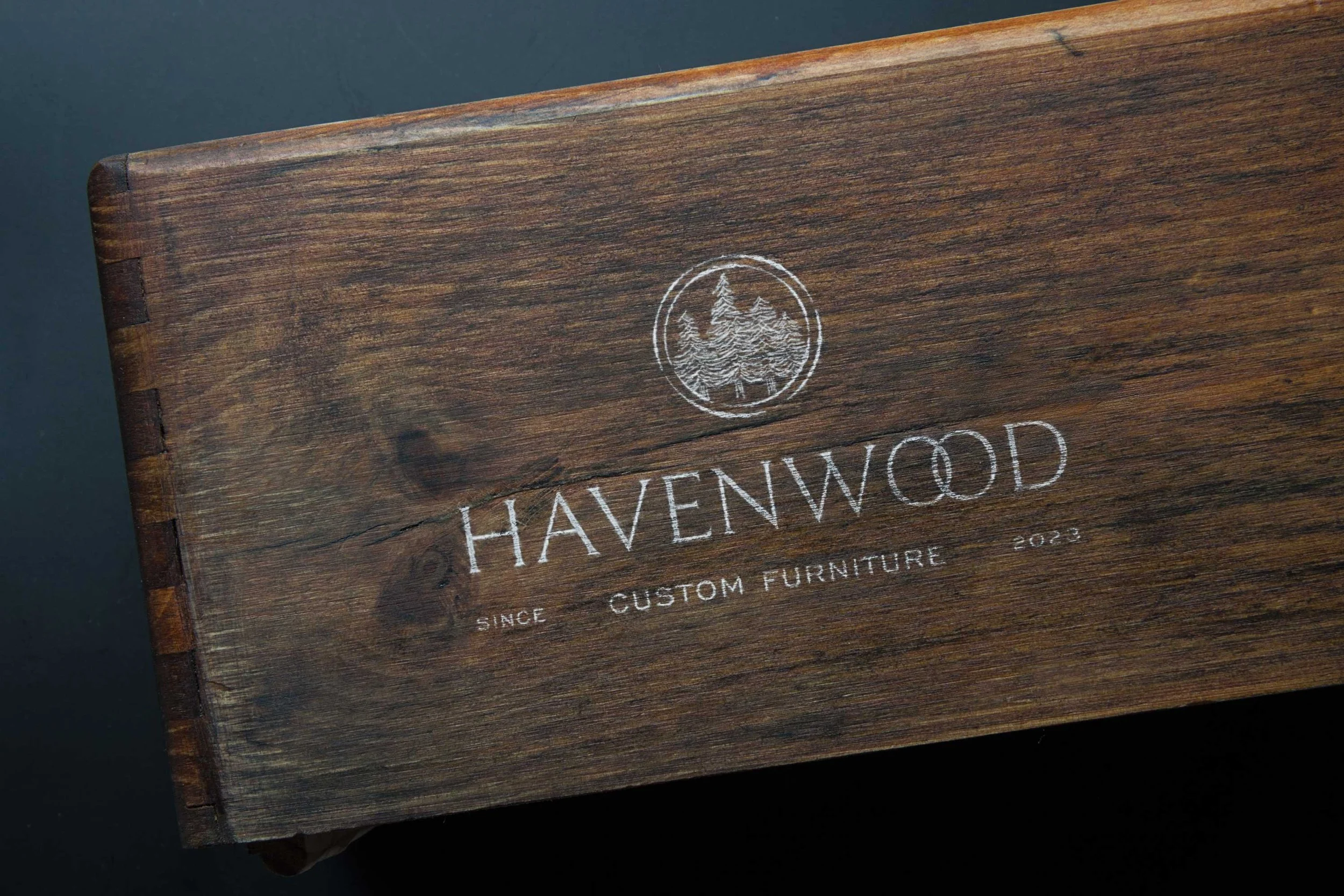 A logo for HavenWood, a custom furniture company since 2023, serving Green Bay, with a background of stylized pine trees in earthy tones.