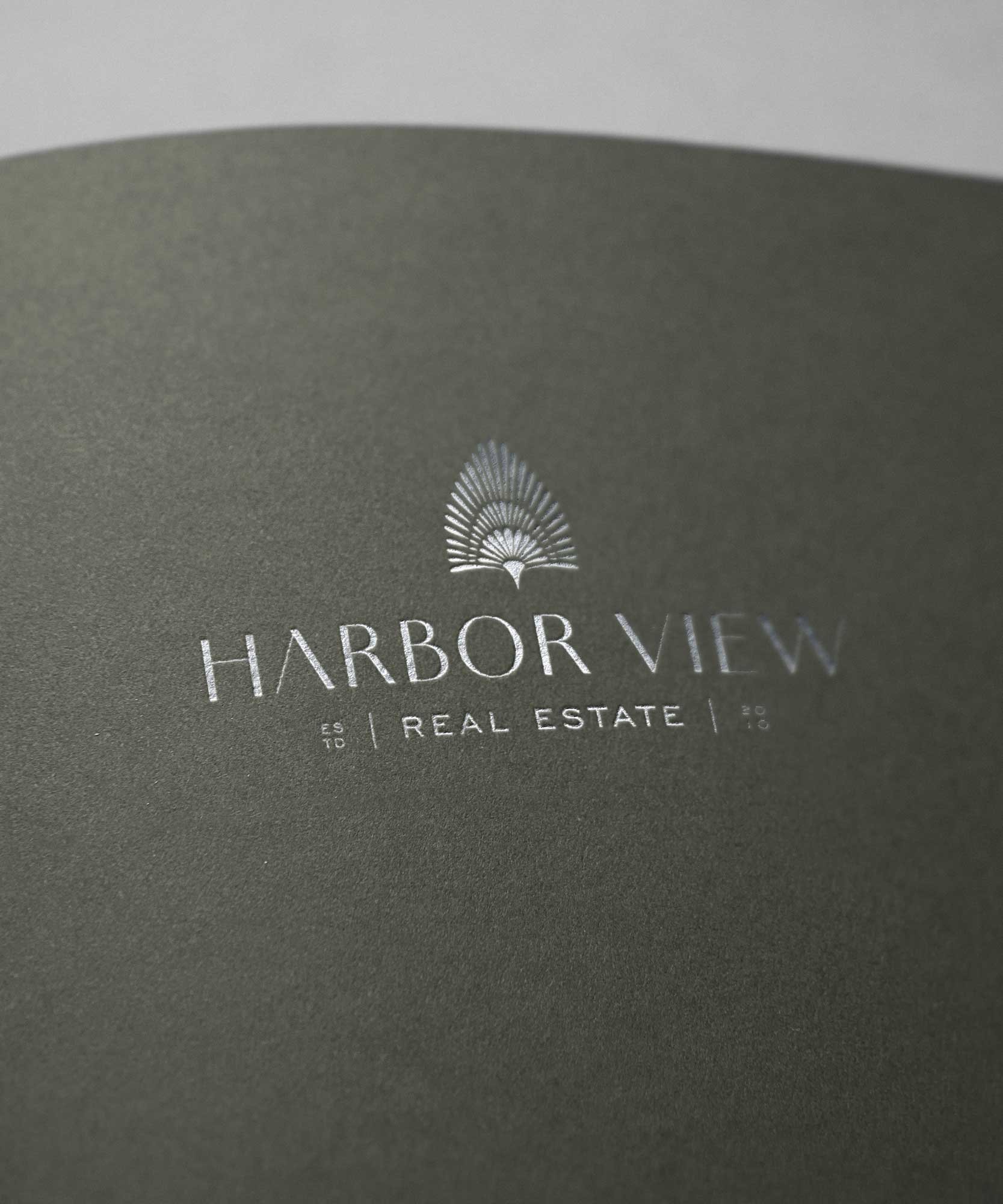 Close-up of a dark green textured surface with a silver embossed logo of a stylized peacock feather and the text 'Harbor View Real Estate'.
