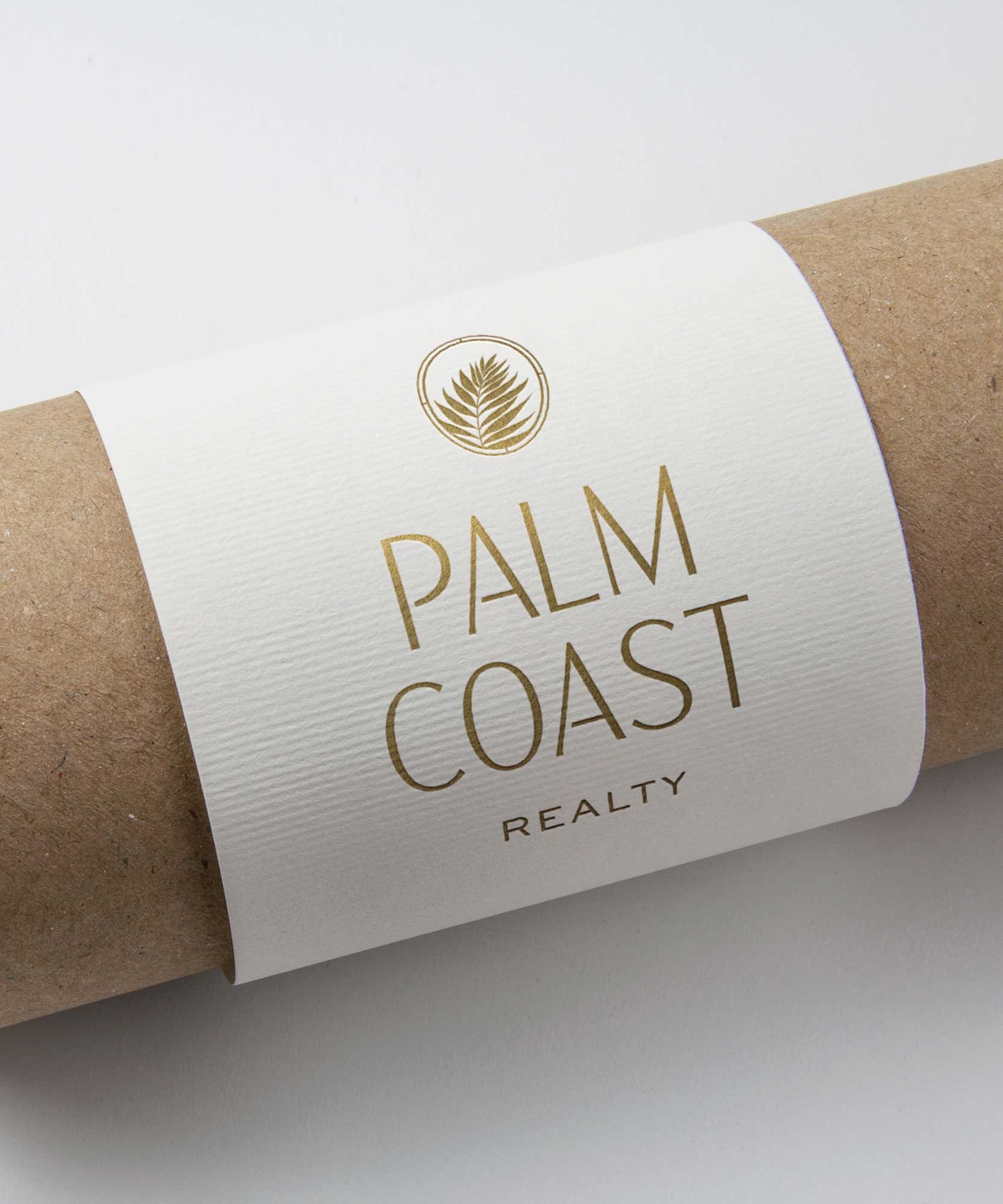 A cylindrical cardboard tube with a white label that has a gold palm leaf logo and gold text reading 'Palm Coast Realty'.