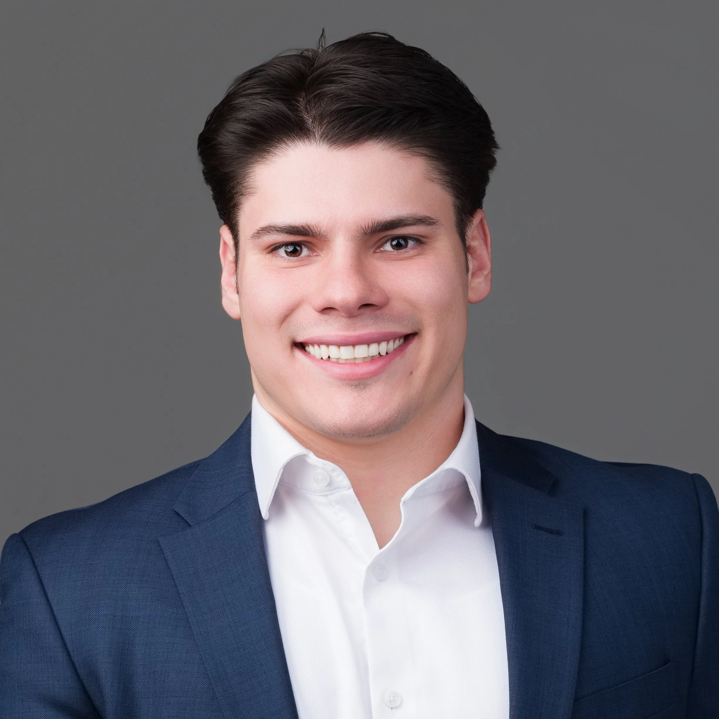 A young man in a navy blue suit, white shirt, and matching navy tie smiling against a white background.