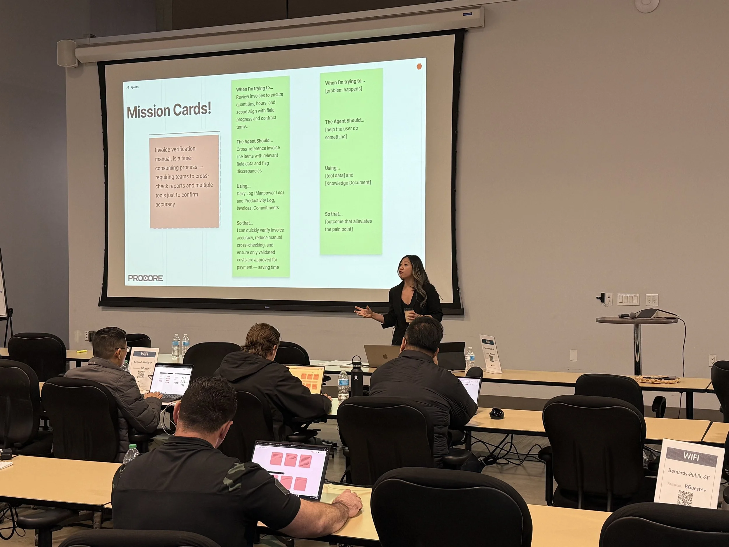 A woman giving a presentation in a conference room with a large screen displaying a presentation slide titled 'Mission Cards!' The slide contains bullet points about invoice verification and agent tasks, with a group of attendees seated and working on laptops at tables in front of her.
