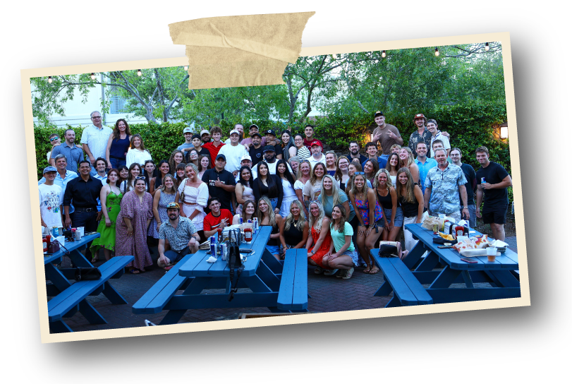 A large group of people gathered outdoors in a backyard, standing and sitting in front of trees and bushes, with picnic tables holding food and drinks, during daytime or early evening.
