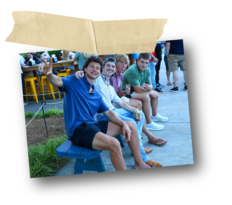 Four young men sitting on a bench outdoors, smiling and waving at the camera, with a crowded event in the background.