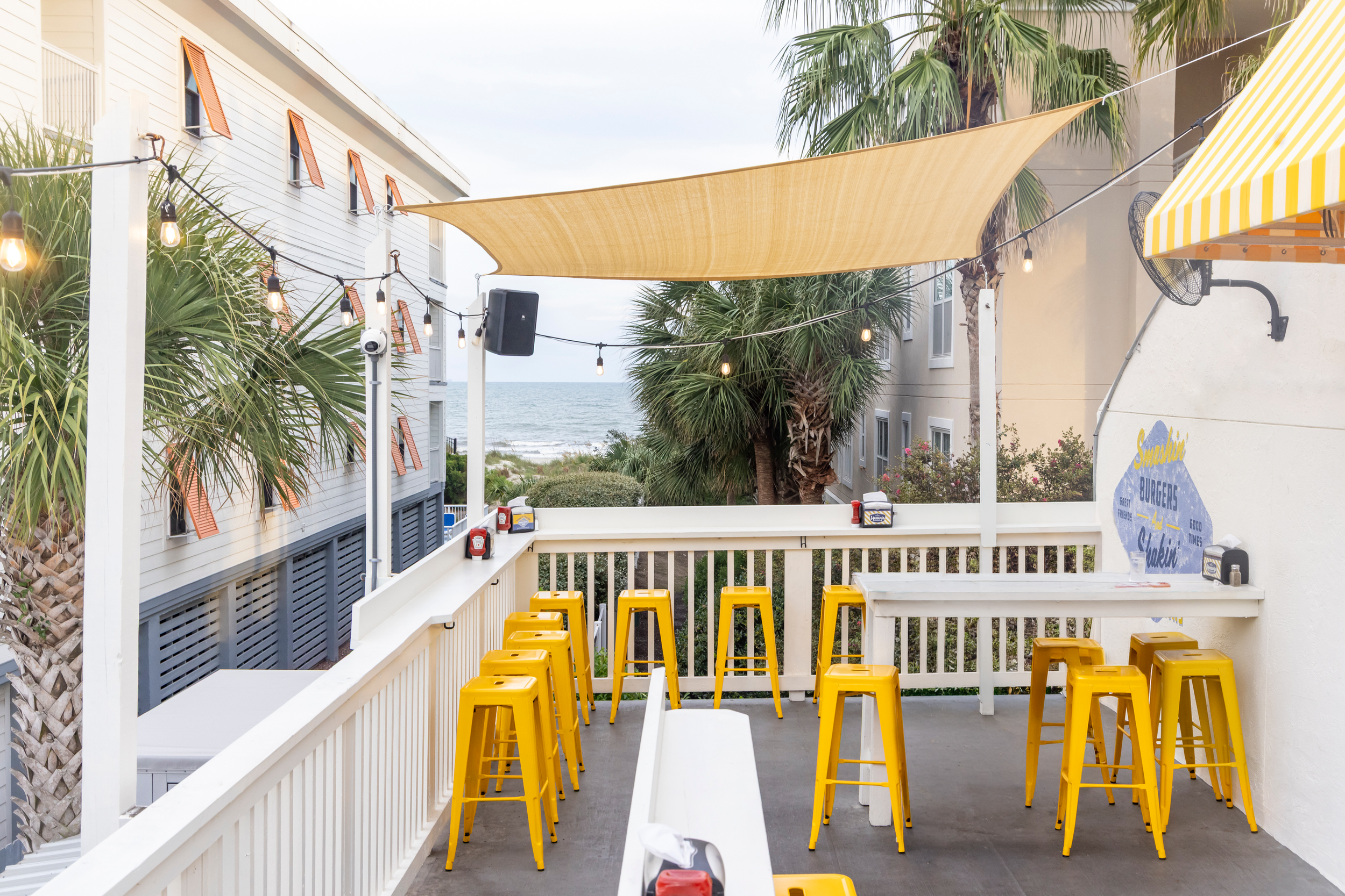 A photo of the upstairs patio of Heavy's at Isle of Palms, showing the ocean and beach in the background, and yellow stools on the patio in the foreground.
