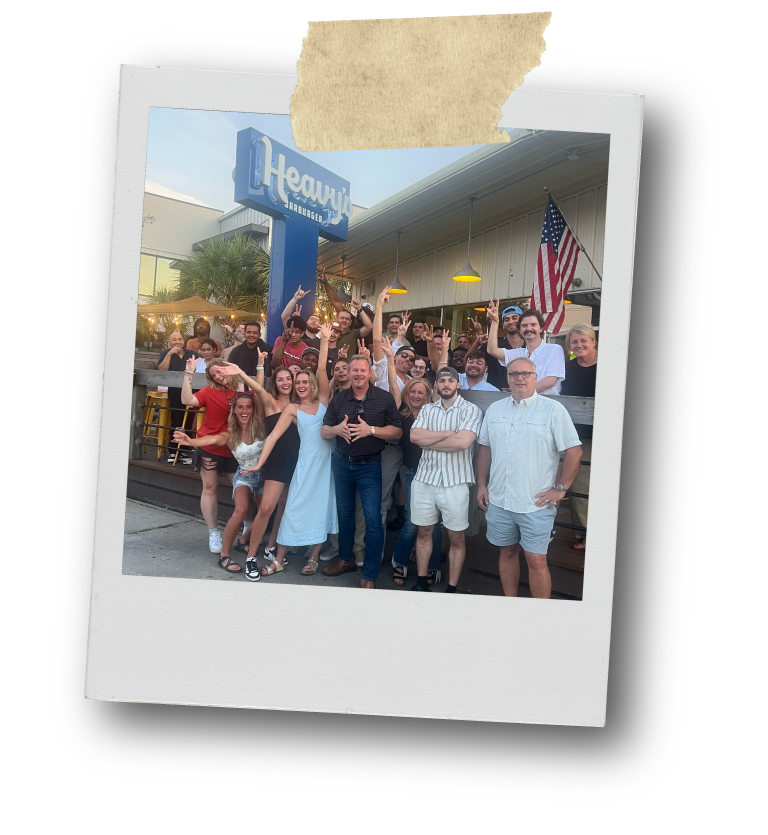 Group of people posing outside a restaurant with a sign that reads 'Heavy Burger' and an American flag, some making peace signs or raising their arms in celebration.