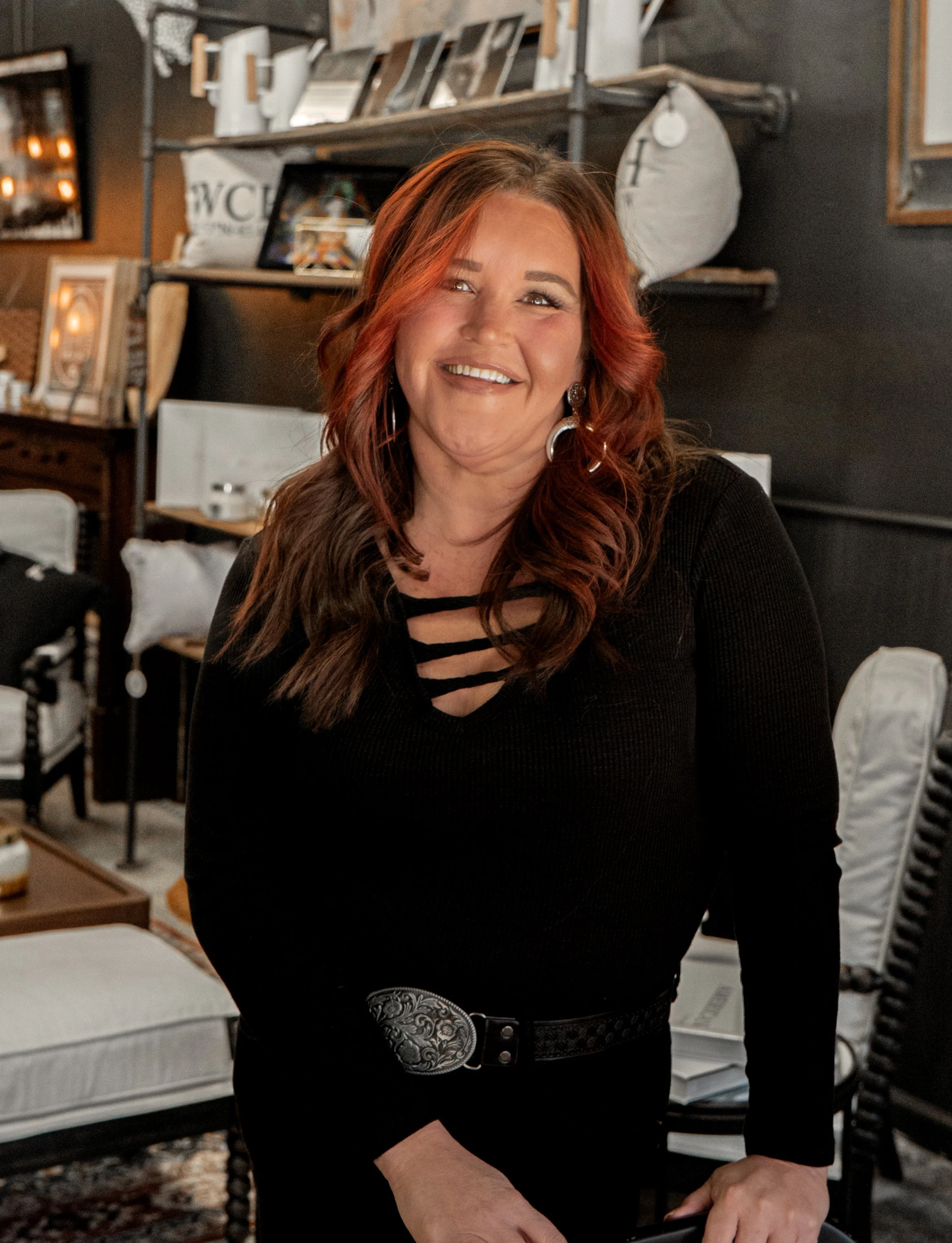 A woman with long reddish-brown hair smiling in an indoor setting with shelves filled with picture frames and decor items in the background.
