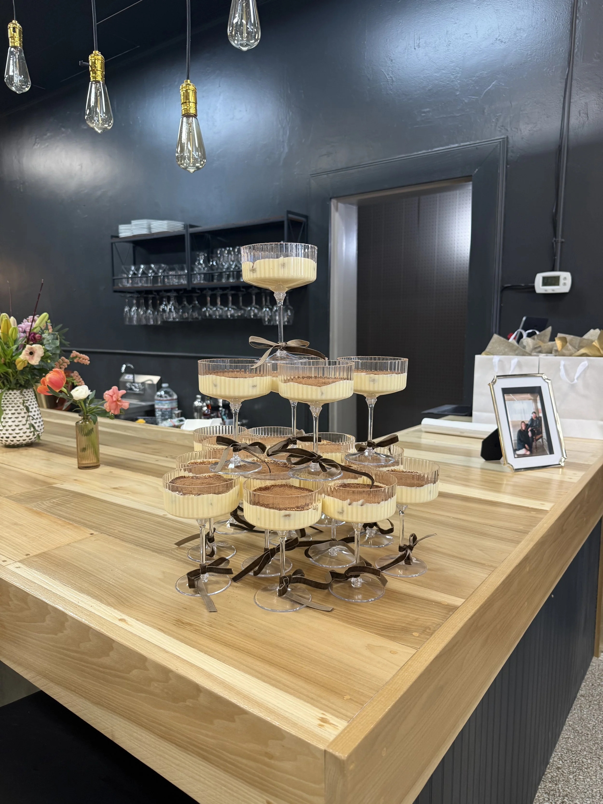 Stacked desserts on wine glasses on a wooden table, with flowers and framed photograph nearby in a modern interior setting.