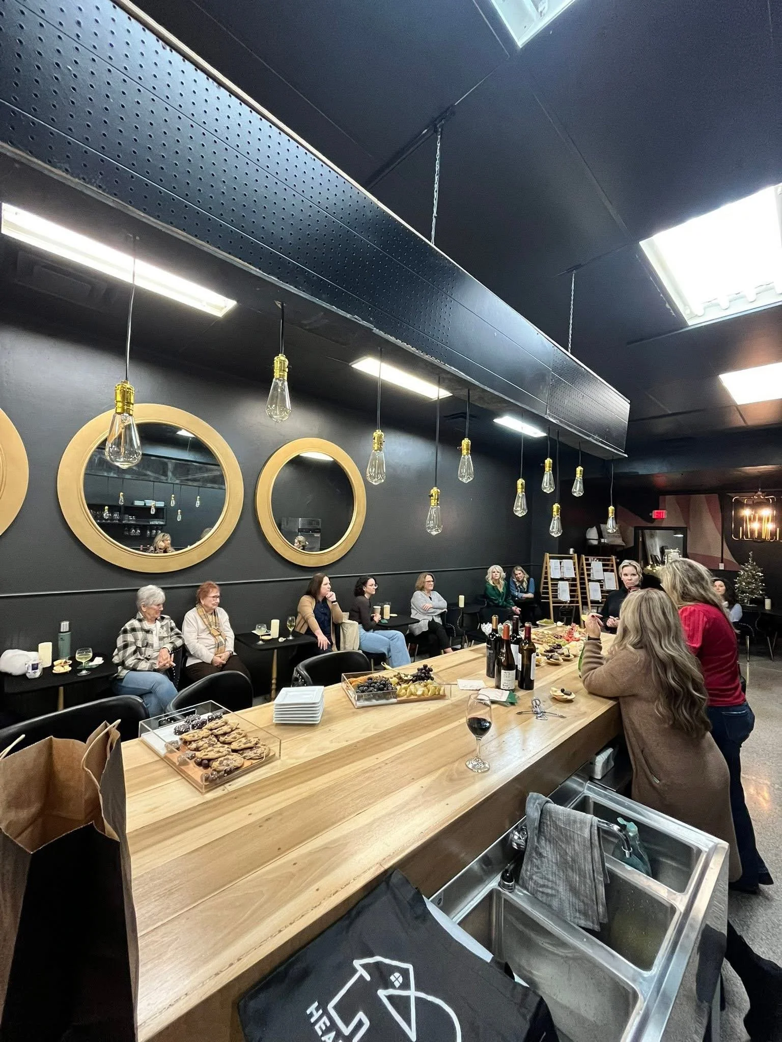 A group of people sitting at a long wooden table in a room decorated with black walls and circular mirrors, enjoying snacks and drinks. The table has cookies and wine. The room has modern hanging lights and a small Christmas tree in the corner.