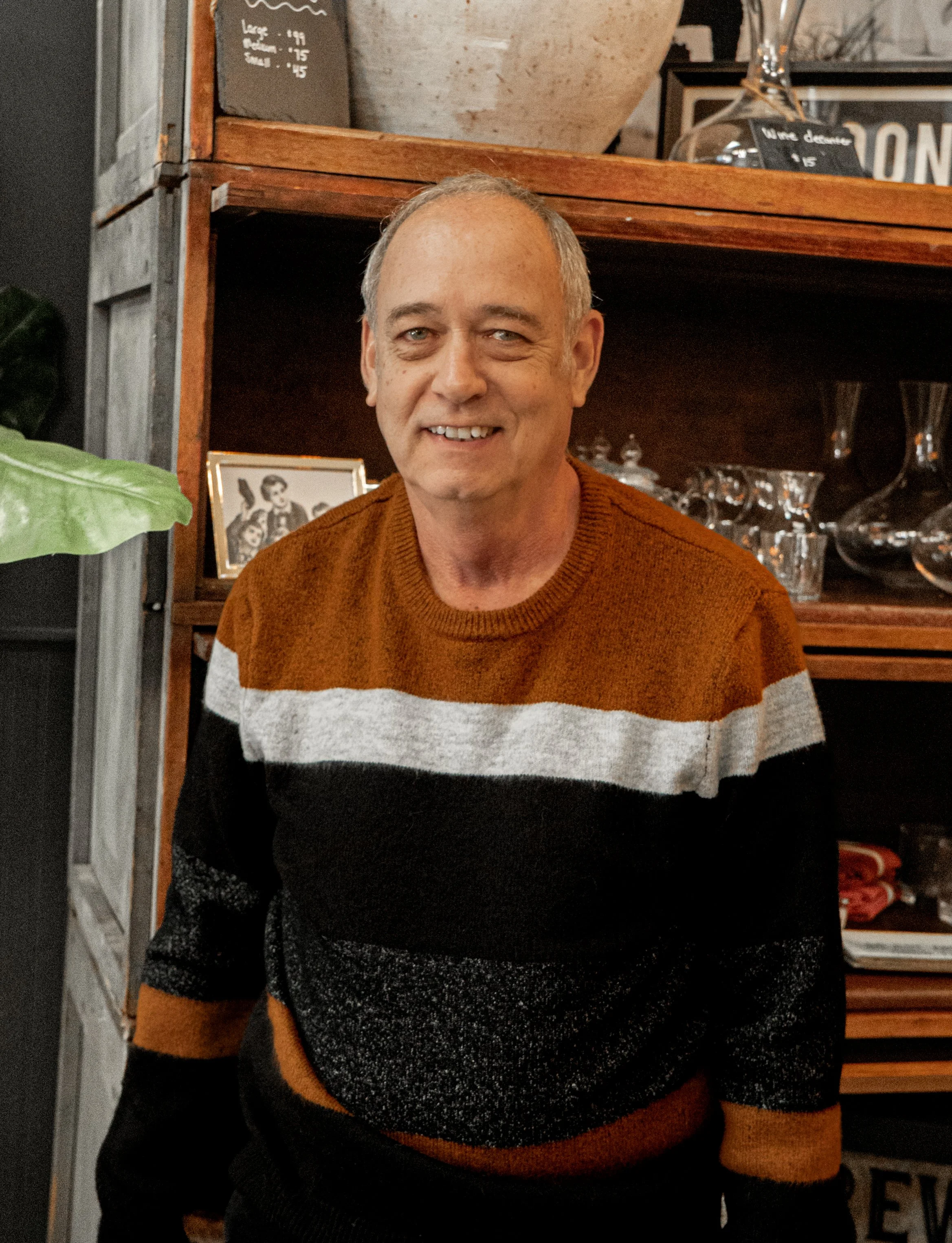A smiling man wearing a brown, black, and white striped sweater, sitting in a cozy cafe with wooden shelves, glasses, and framed photos in the background.
