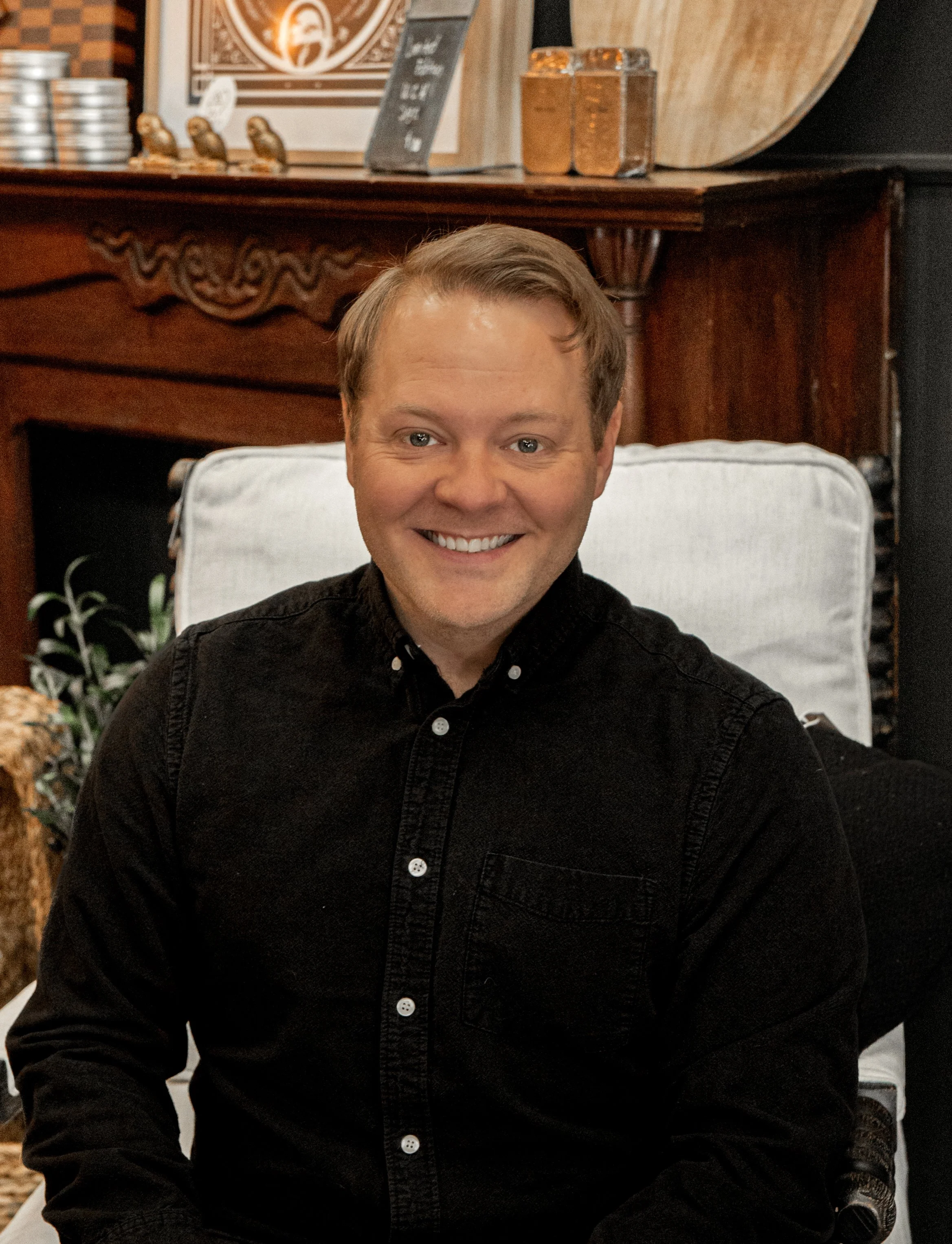 A smiling man with blond hair sitting on a white chair in a cozy, decorated indoor space with wood accents and a chalkboard.