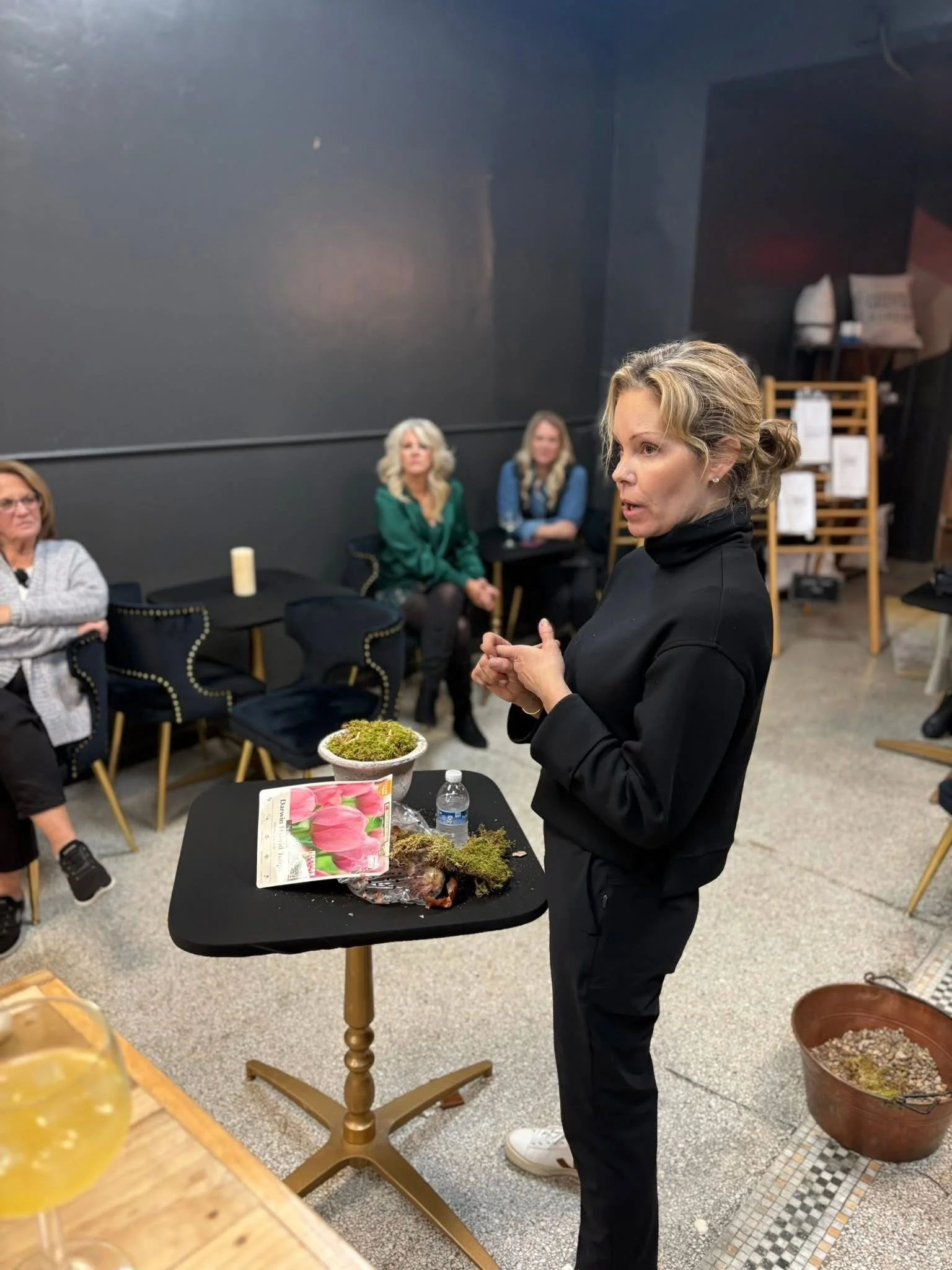 A woman standing at a table with a plant and gardening supplies, speaking to a seated group of women in a room with dark walls, some on chairs and some standing.