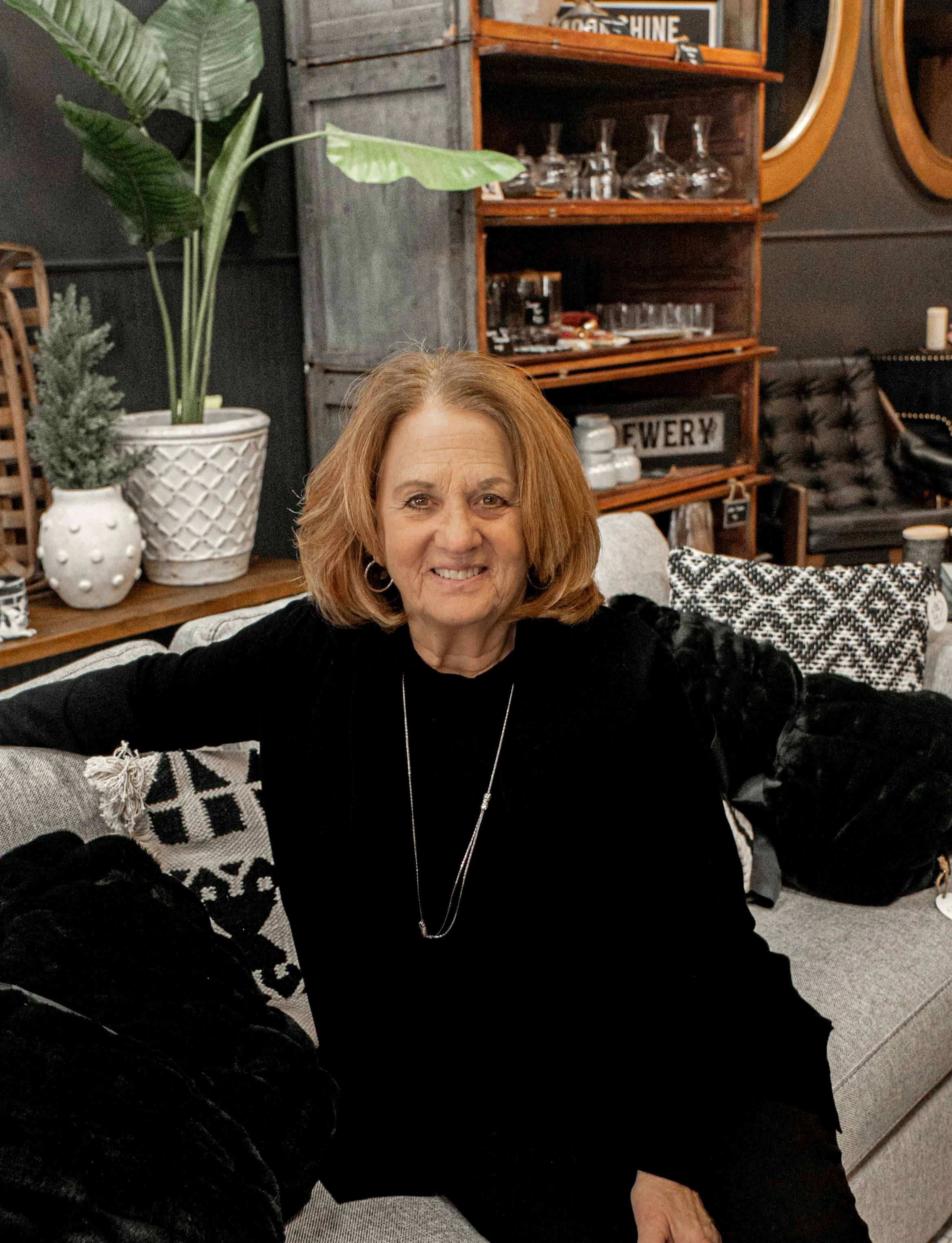 A woman with shoulder-length red hair smiling, sitting on a black and white patterned pillow on a gray sofa. Behind her are decorative padded pillows, a large potted plant, and a wooden shelving unit with glassware and signs in a cozy, stylish living room.